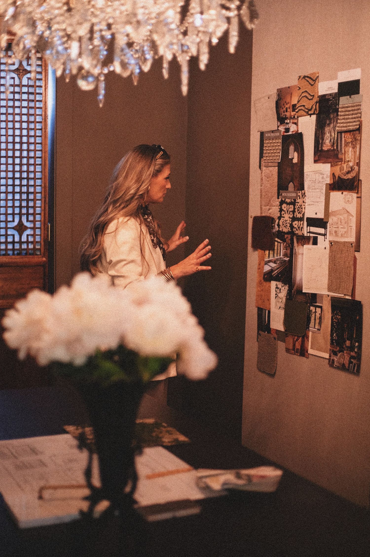 Woman gesturing at a mood board on a wall, with white flowers and a crystal chandelier in a warmly lit room.