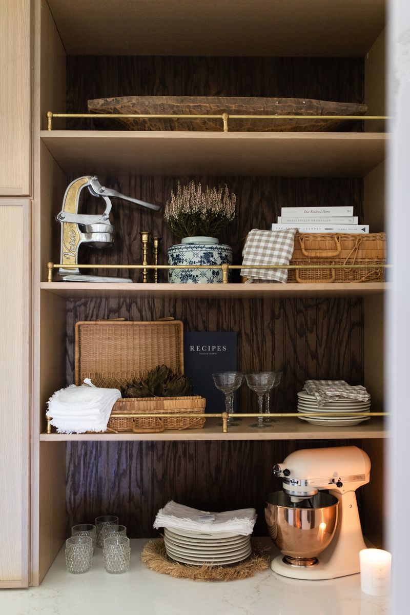 Styled kitchen shelves with wicker baskets, wine glasses, stacked plates, a KitchenAid mixer, and decorative items on wood-backed shelving.