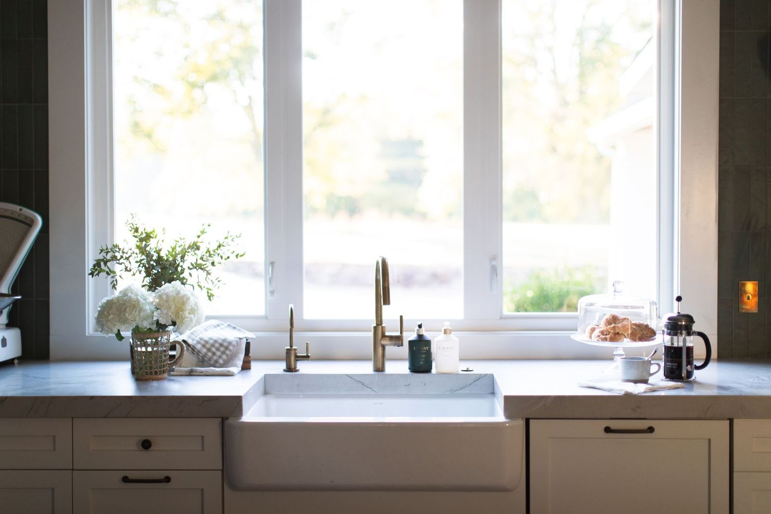 Bright farmhouse kitchen sink with gold faucet, white cabinets, flowers, and a large window letting in natural light.