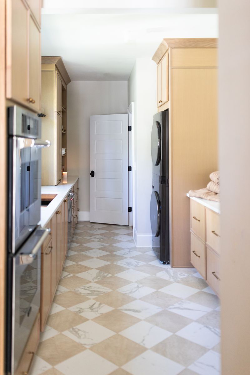 A narrow kitchen hallway with checkered marble floors, wooden cabinetry, stainless steel ovens, and a stacked washer-dryer unit.