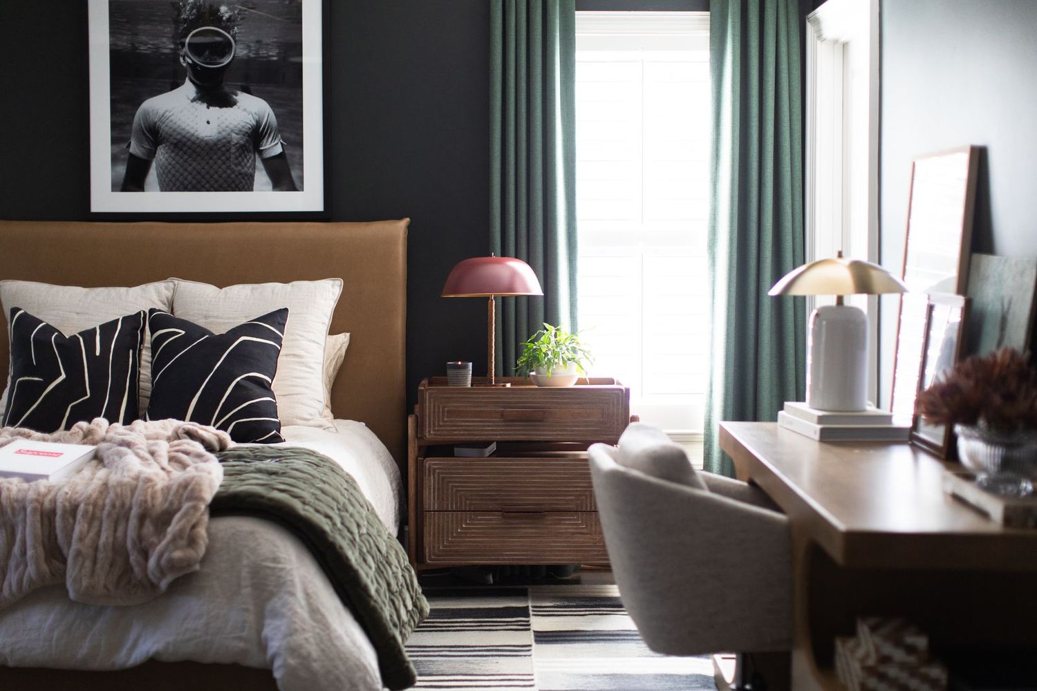 Cozy bedroom with tan upholstered bed, dark walls, green curtains, wooden nightstand, and stylish lamps on a desk.