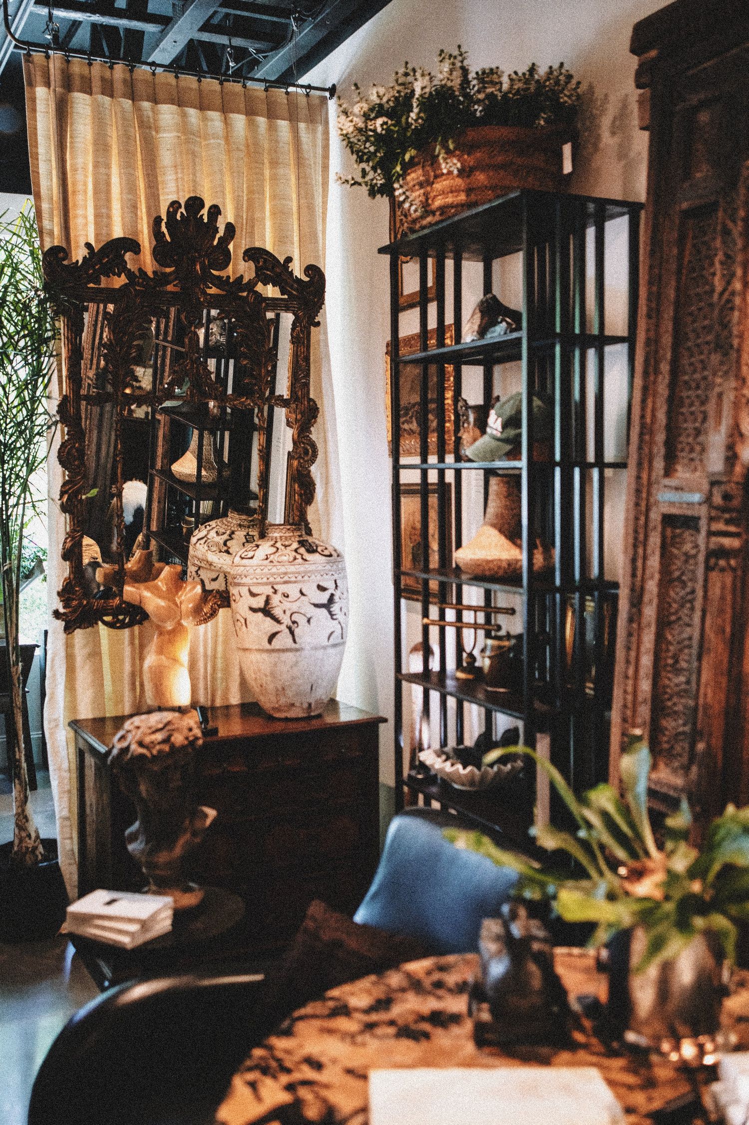 Eclectic home decor shop with ornate wooden mirror, ceramic vases, metal shelving unit, and lush greenery in warm lighting.