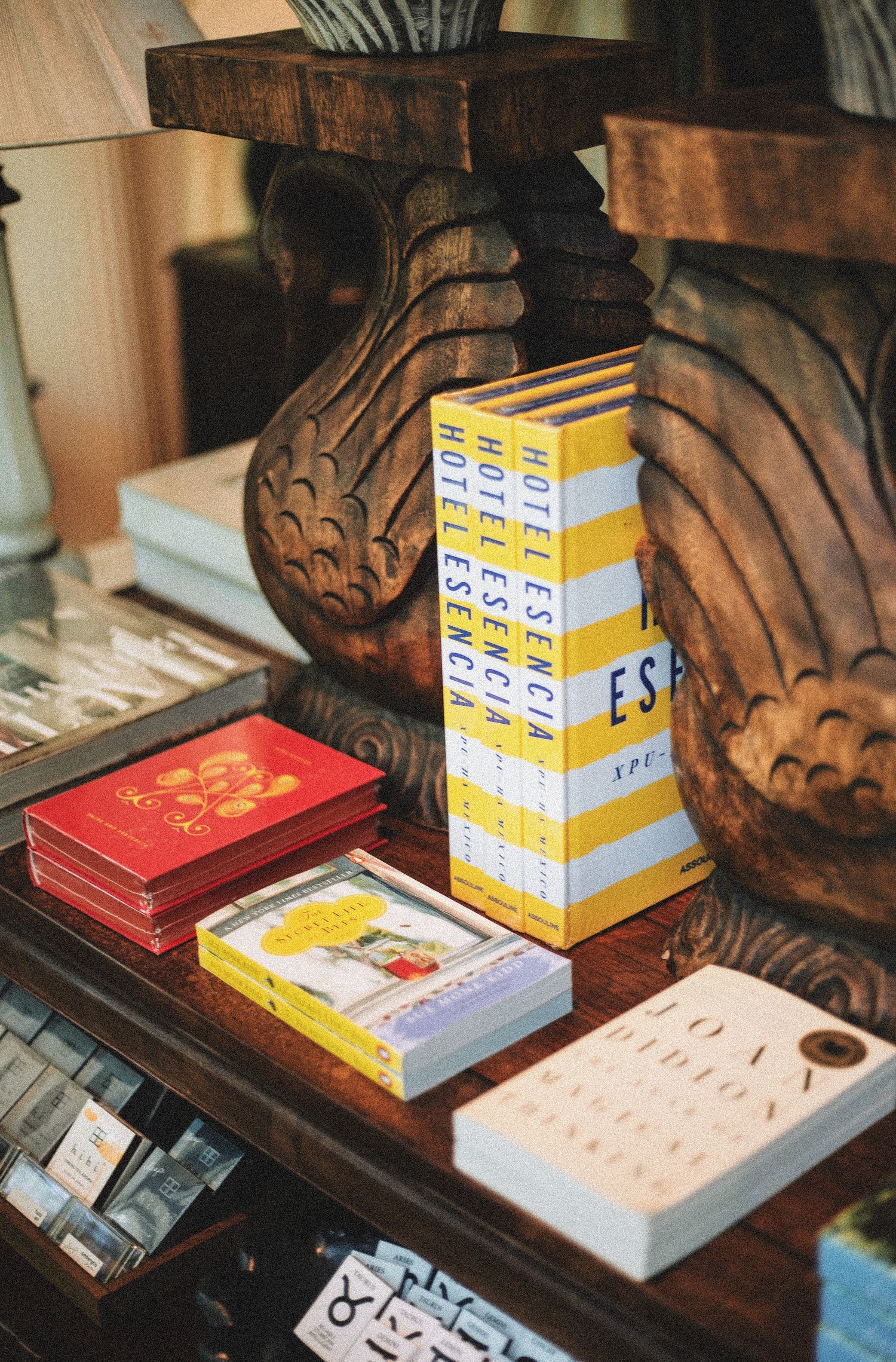 Stack of books including Hotel Esencia editions displayed on ornate wooden shelf with carved decor.