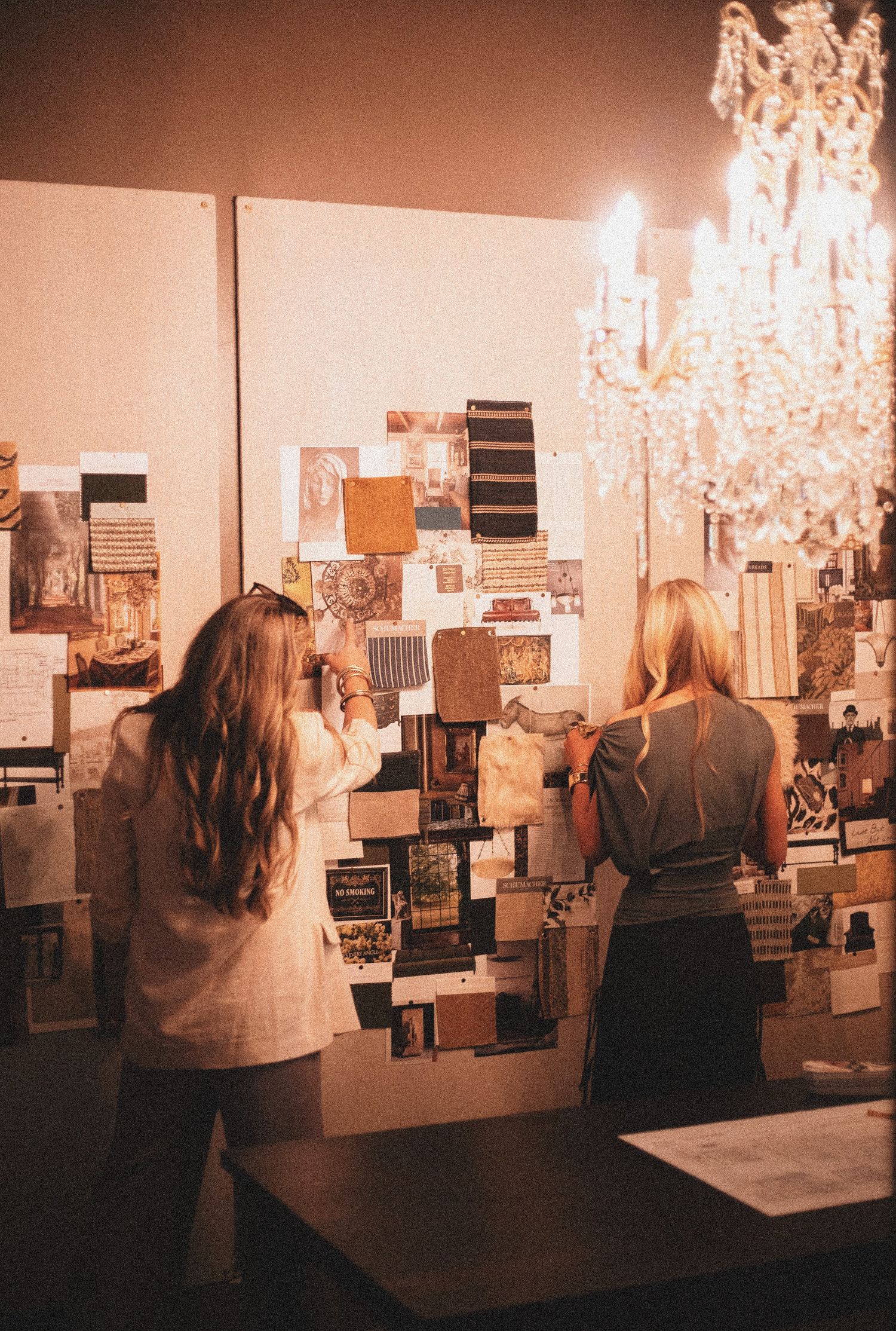 Two women reviewing a mood board with fabric swatches and images under a crystal chandelier in a warmly lit studio.