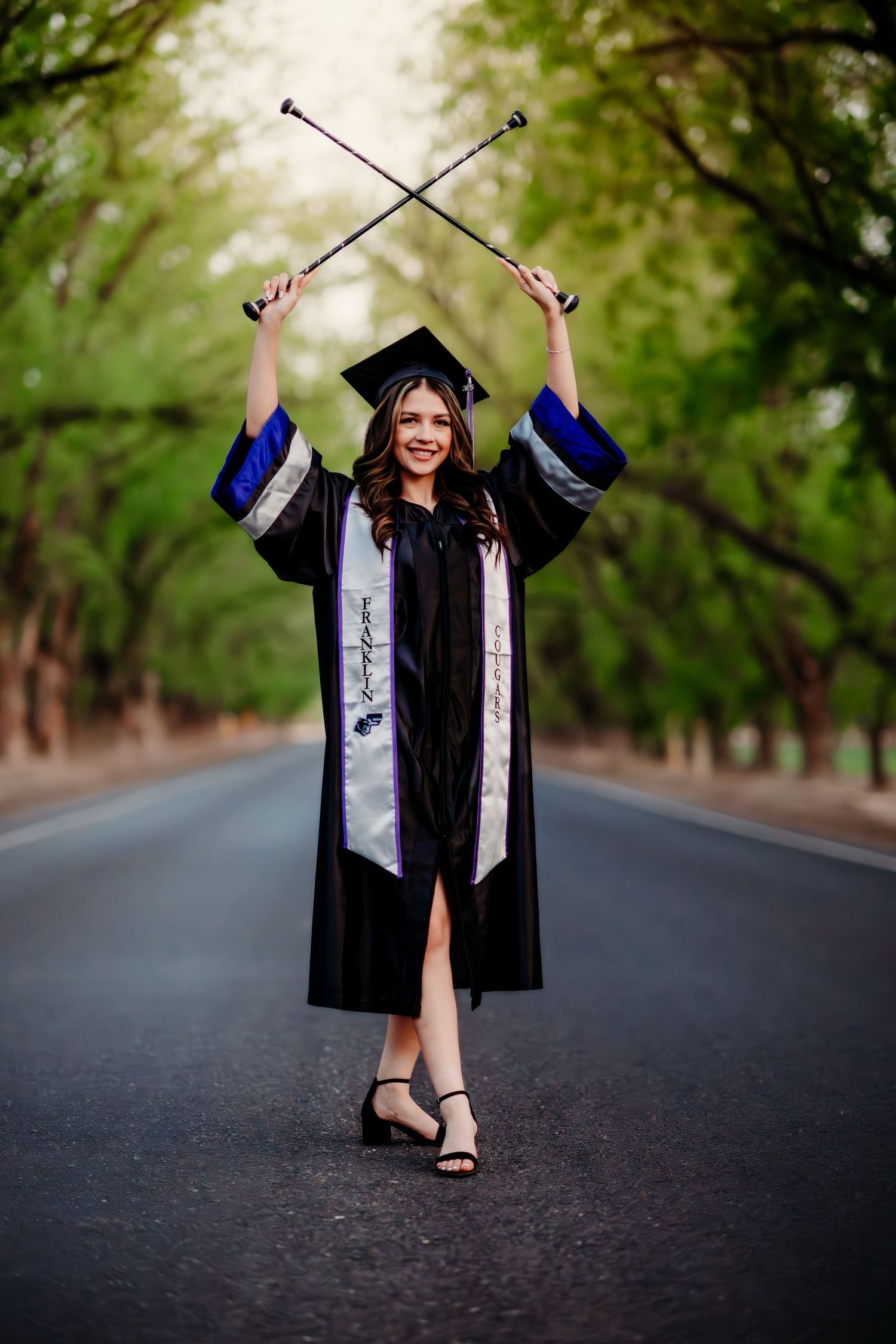 Graduate in black cap and gown poses on empty tree-lined road during outdoor graduation photoshoot session.
