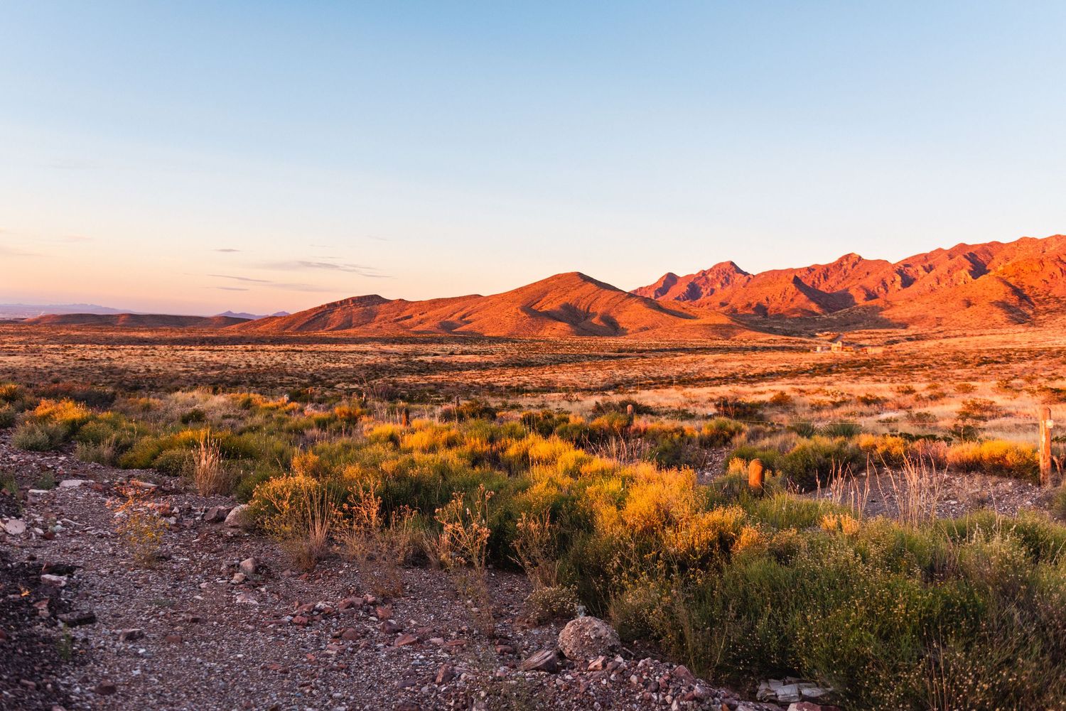 Sunset illuminates red mountains across a desert landscape with scattered brush.