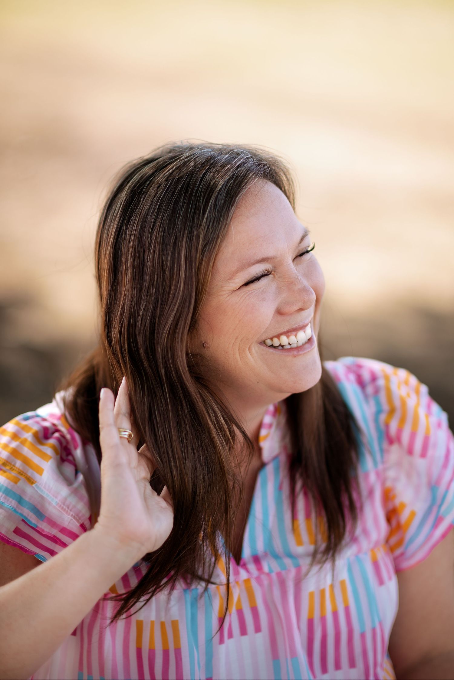 A joyful person wearing a colorful tie-dye shirt laughs while touching their hair during a sunset photoshoot.