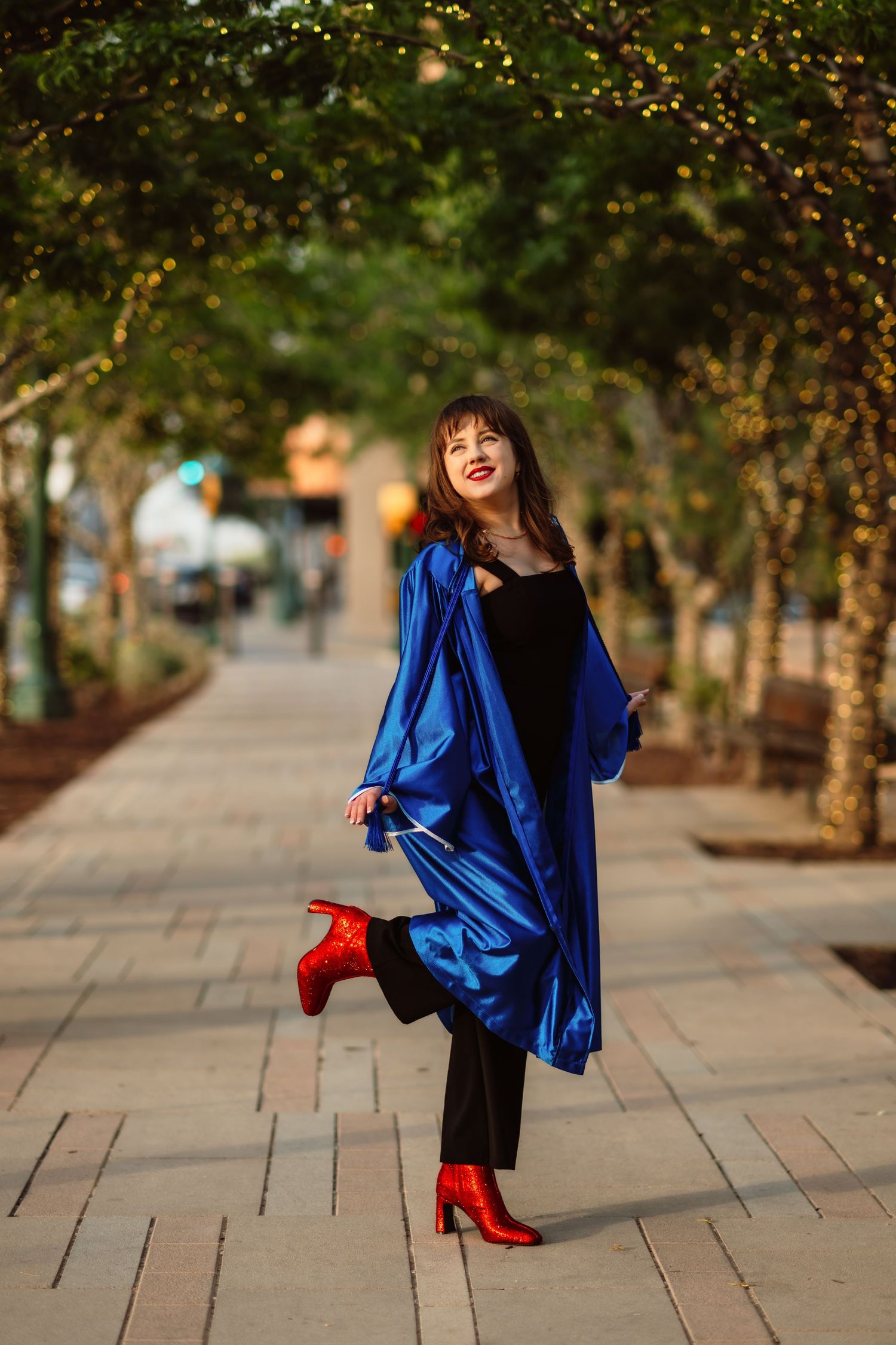Woman in flowing blue coat and red heels poses playfully on urban sidewalk lined with twinkling trees.
