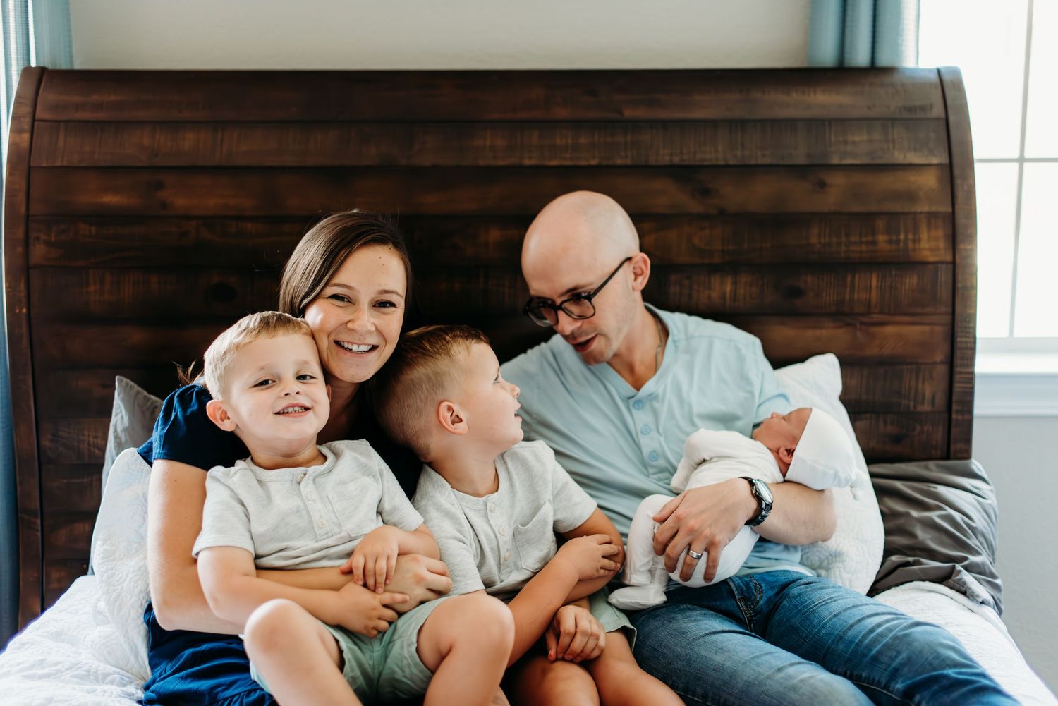 Family relaxing together on brown wooden headboard bed during intimate home photoshoot.