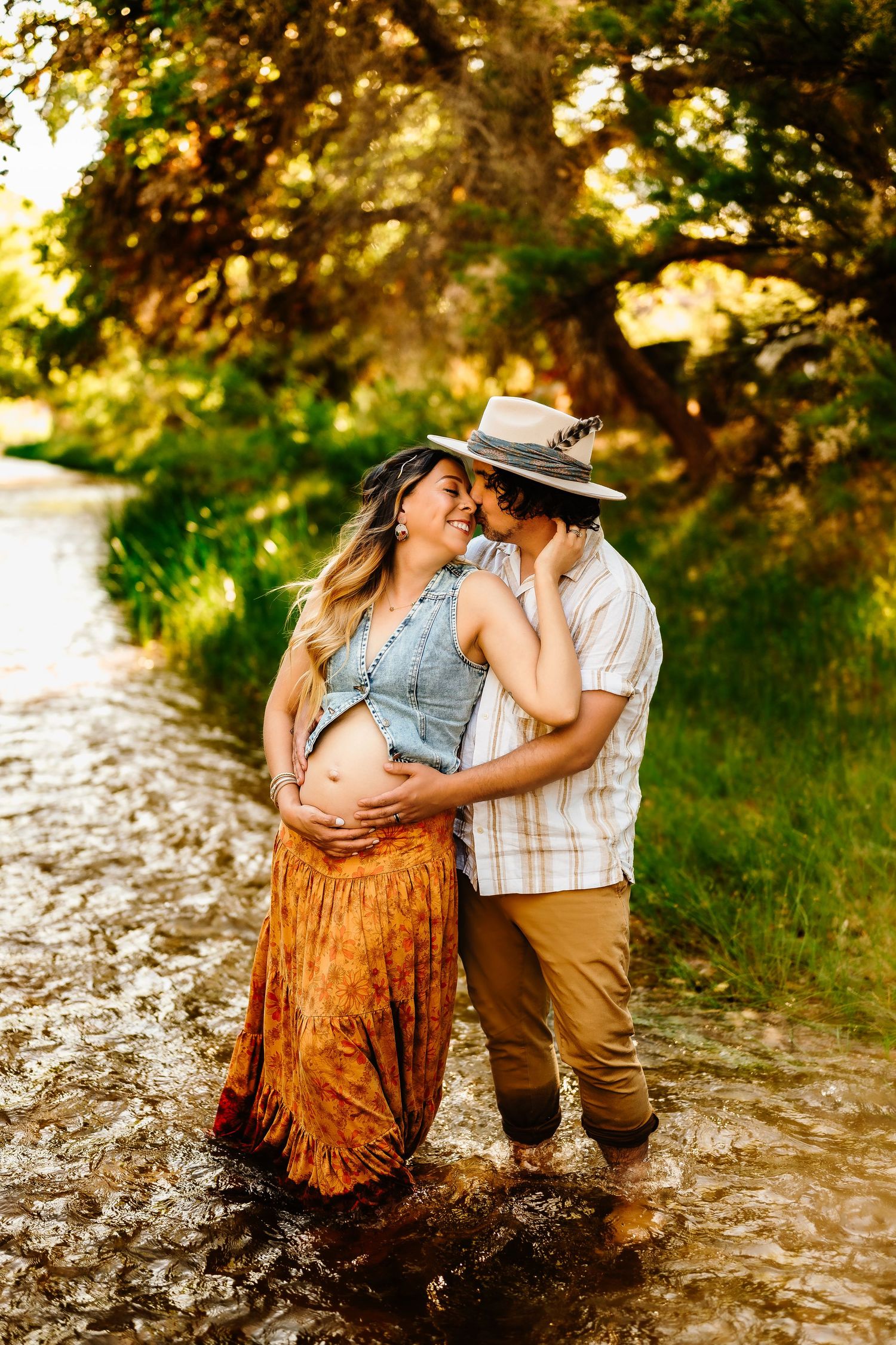 A couple embraces on a rustic path surrounded by golden sunlit foliage during a maternity photoshoot.