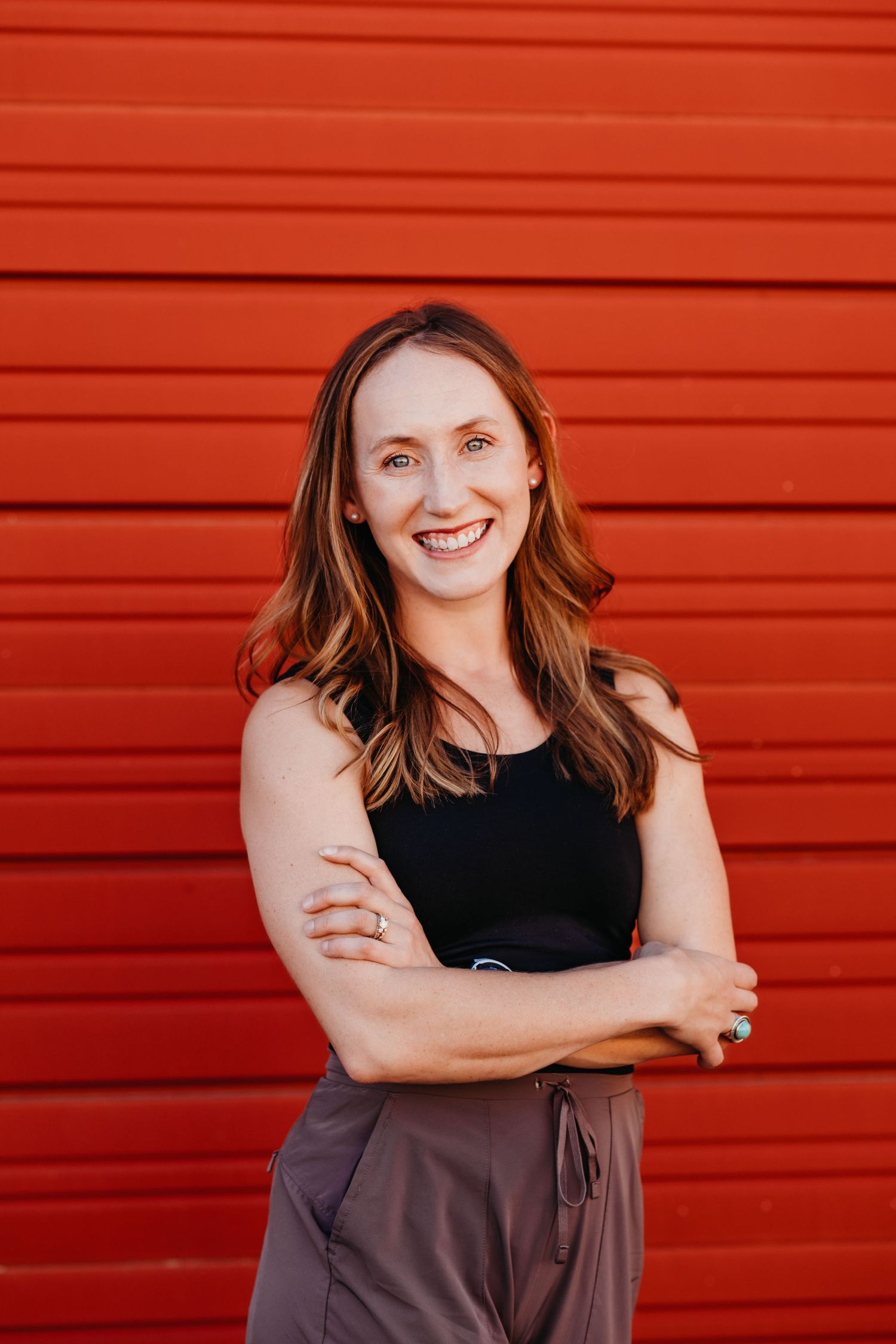 A person in a black sleeveless top and brown pants stands with crossed arms against a bright red wall.