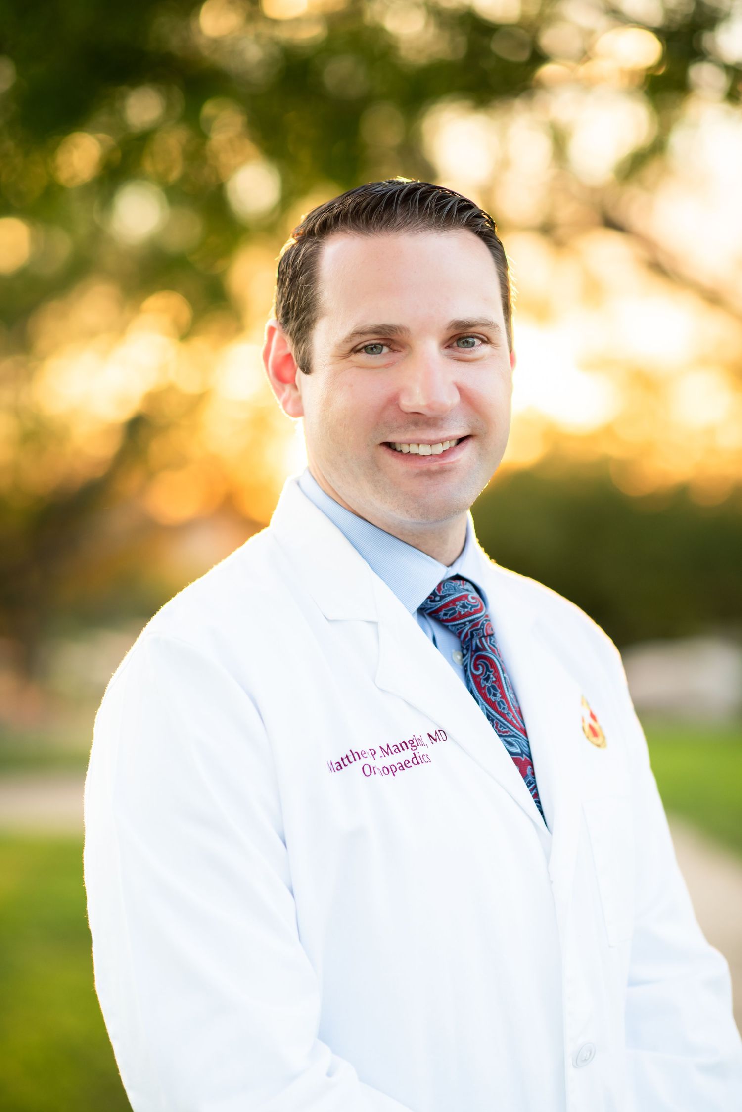 Medical professional in white coat and blue tie poses outdoors with warm sunset lighting in background.