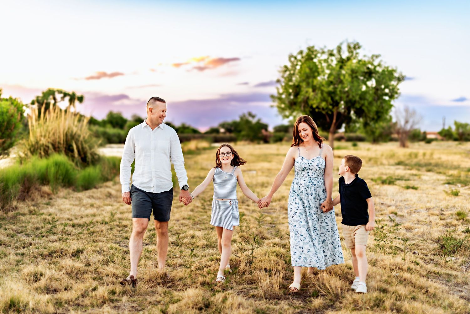 Family of four holding hands walking through a golden field at sunset with trees and colorful sky.