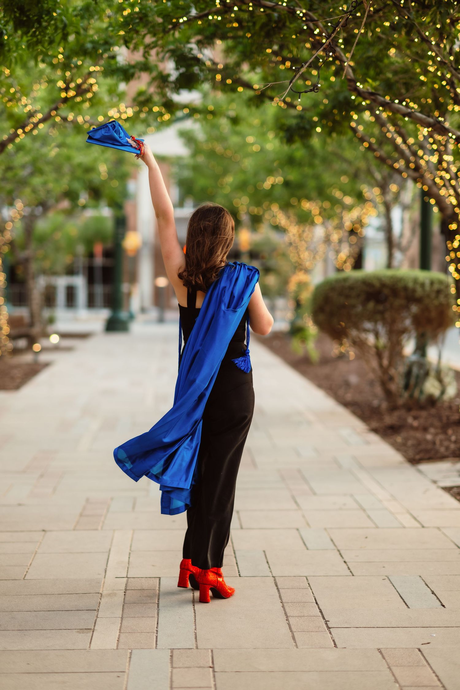 Figure in blue coat and red heels twirls joyfully under illuminated trees on stone pathway.