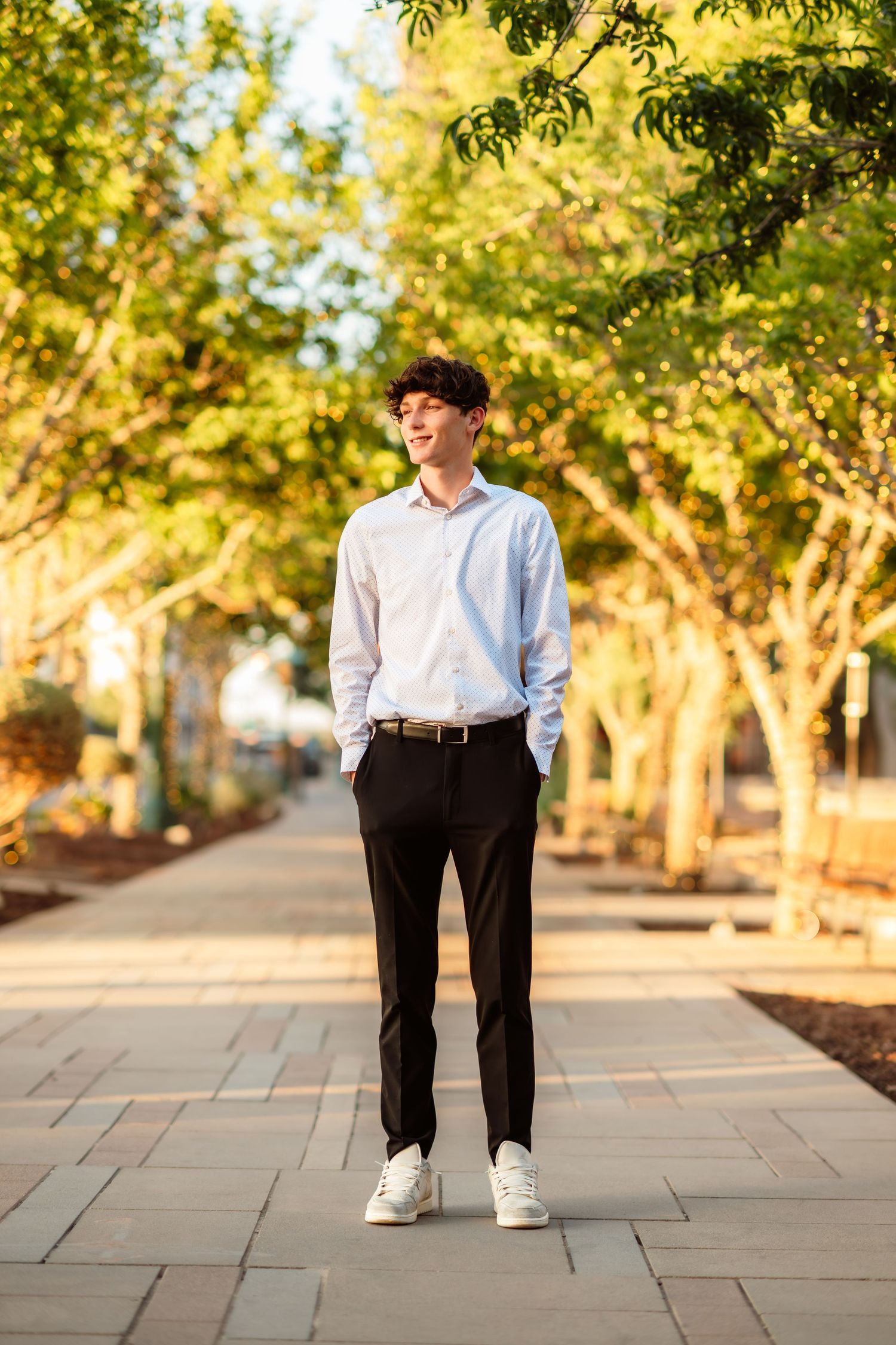 Person in casual white attire walking along a tree-lined stone pathway during golden hour.