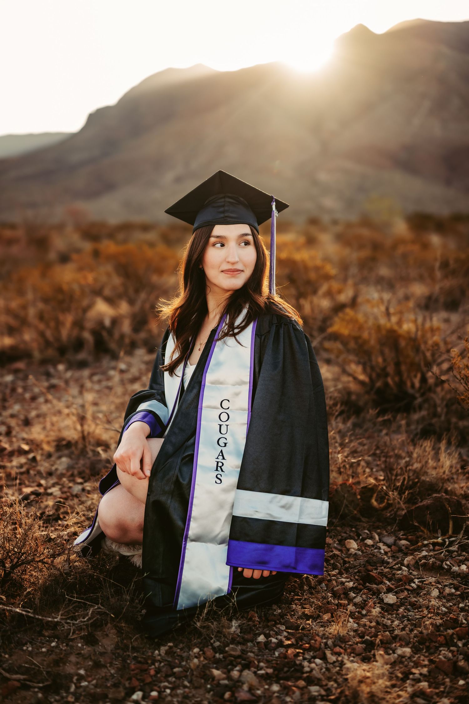 Graduation photoshoot with cap and gown against desert mountain backdrop at sunset.