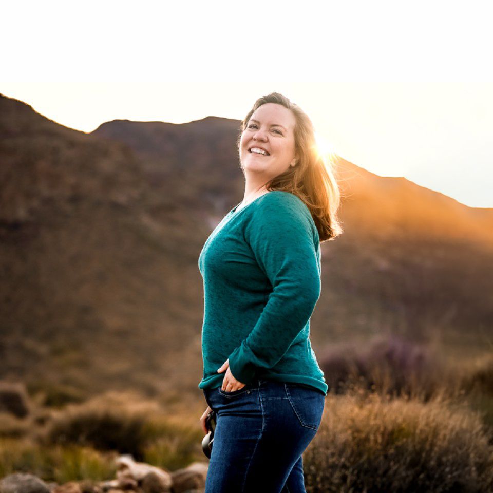 Woman in teal sweater smiles joyfully against desert mountain backdrop during golden hour sunset.