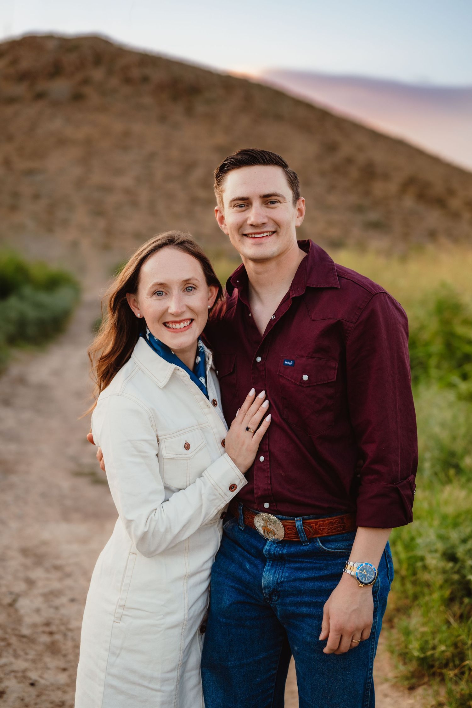 A couple smiles together outdoors wearing burgundy and white casual attire against desert backdrop.