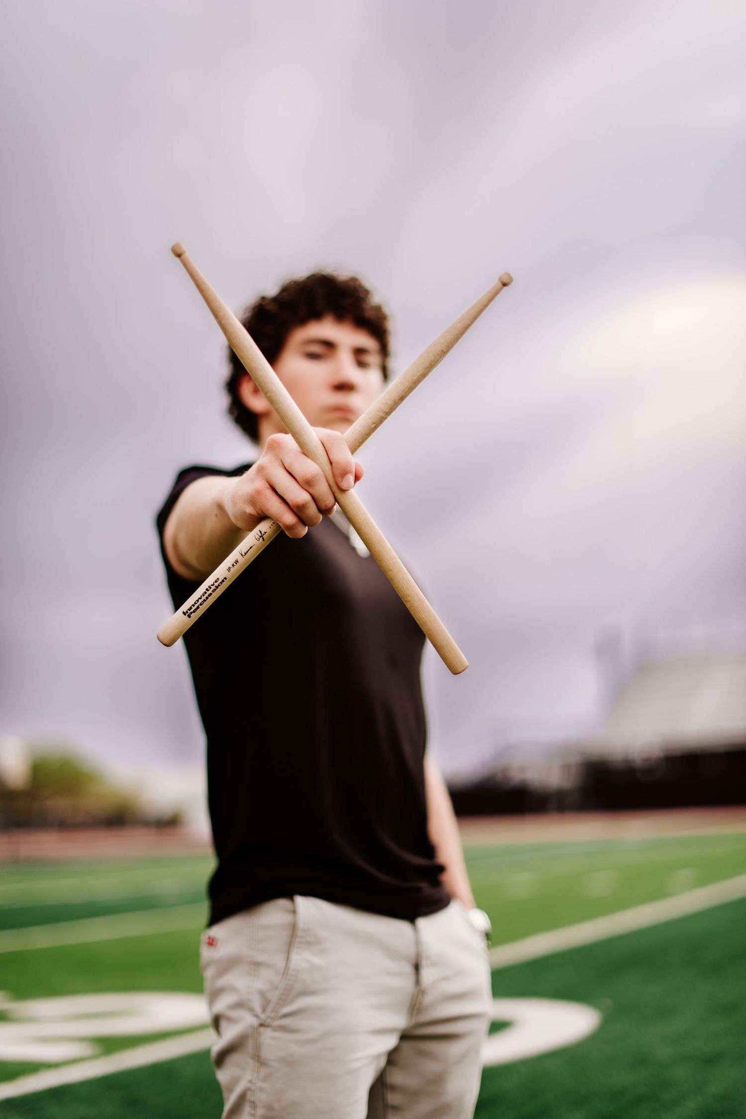 Drumsticks cross in motion against a cloudy sky and green field, creating dynamic percussion performance angles.