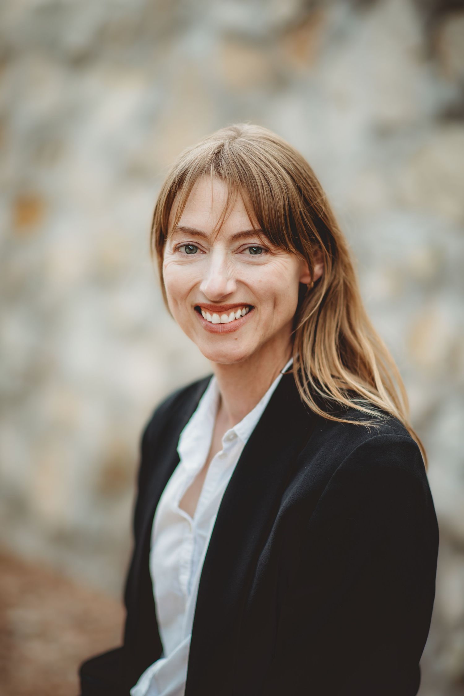 Professional headshot of a person in a black blazer and white shirt smiling against a blurred background.