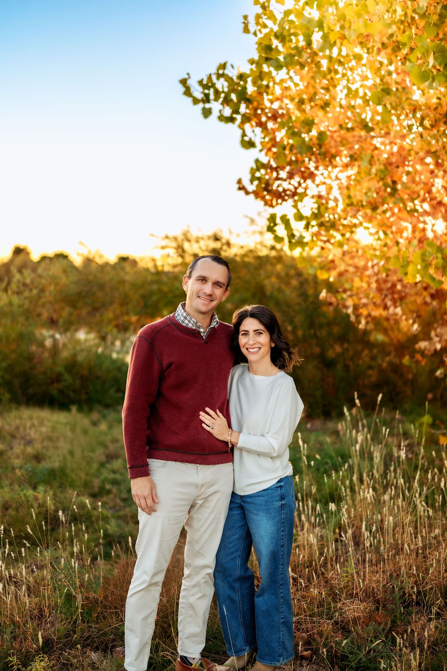 A couple poses together in an autumn field with golden foliage in the background.