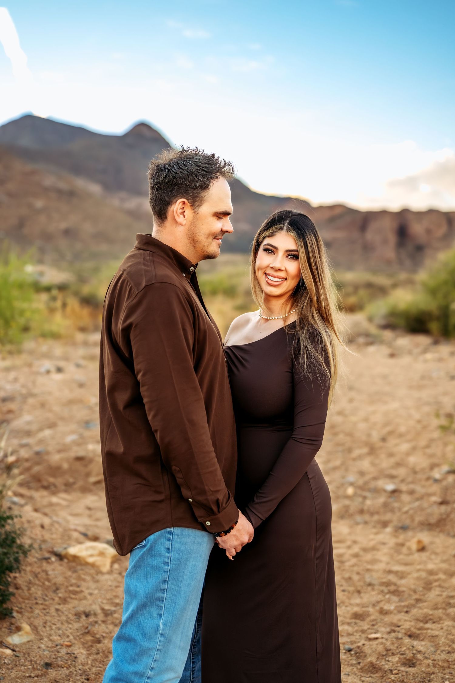 A couple stands close together in a desert setting wearing coordinated brown outfits.