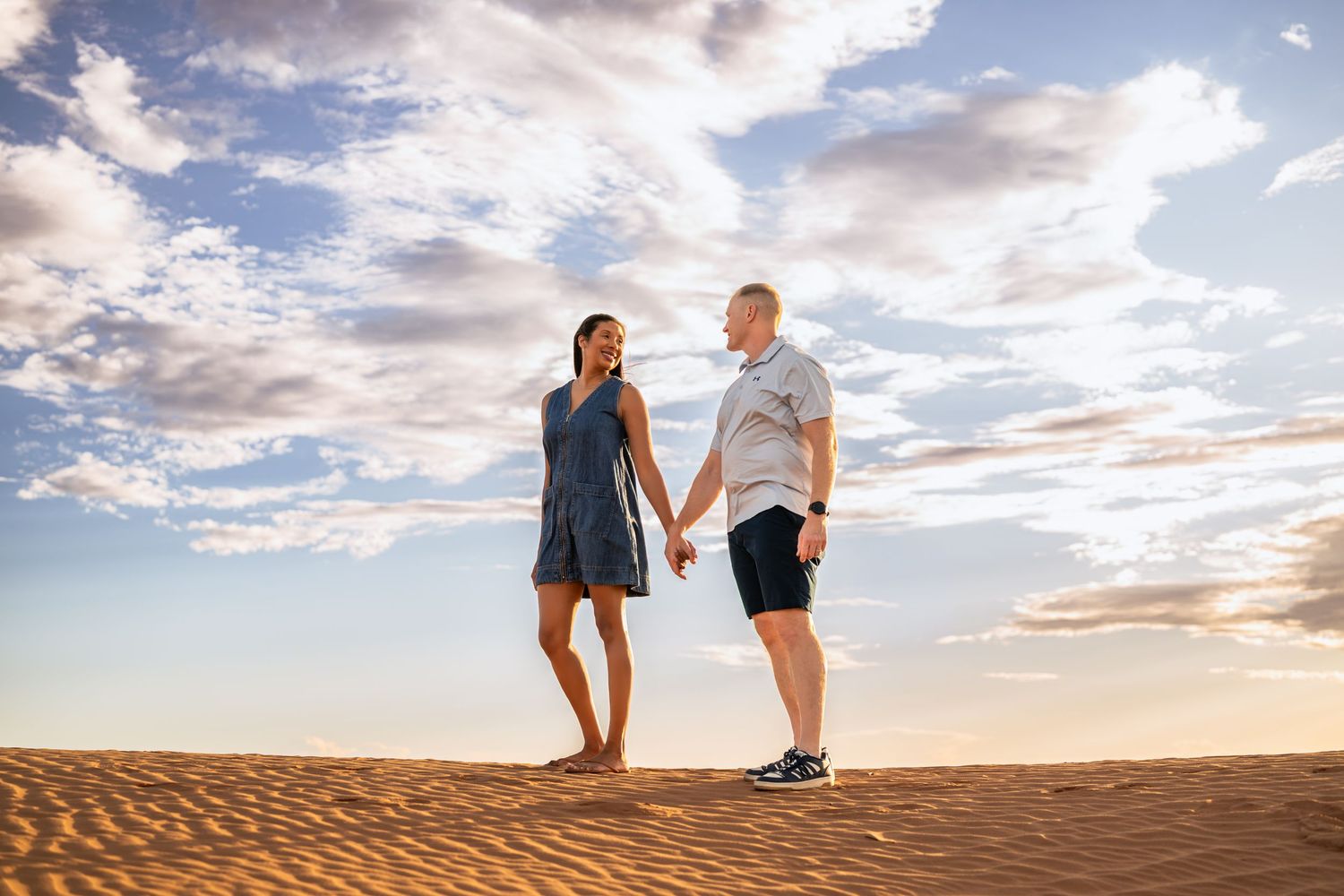 Couple holding hands on sandy desert dunes under a dramatic cloudy sky at golden hour.