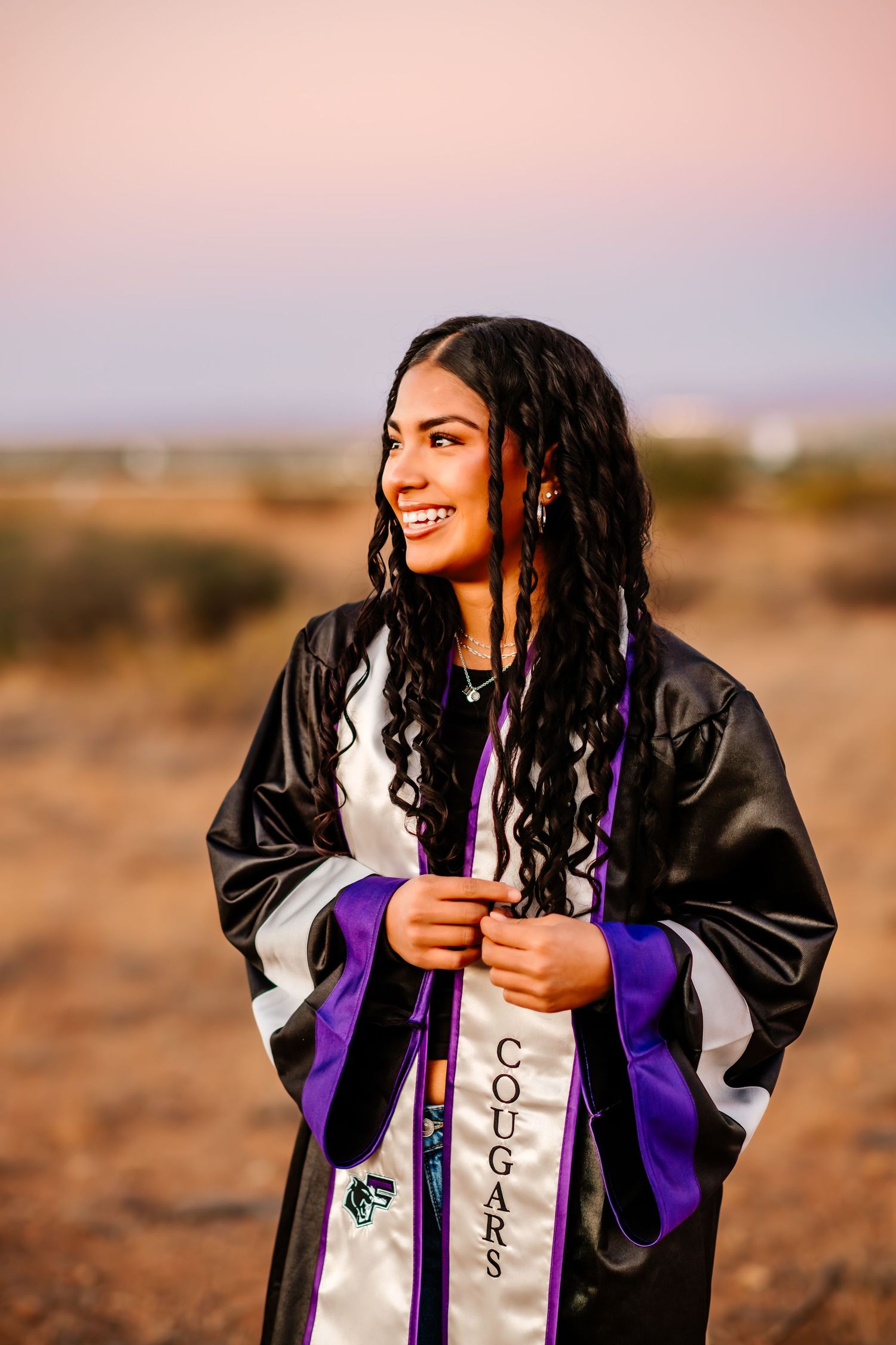 Graduate in black and purple graduation gown smiles against desert sunset.