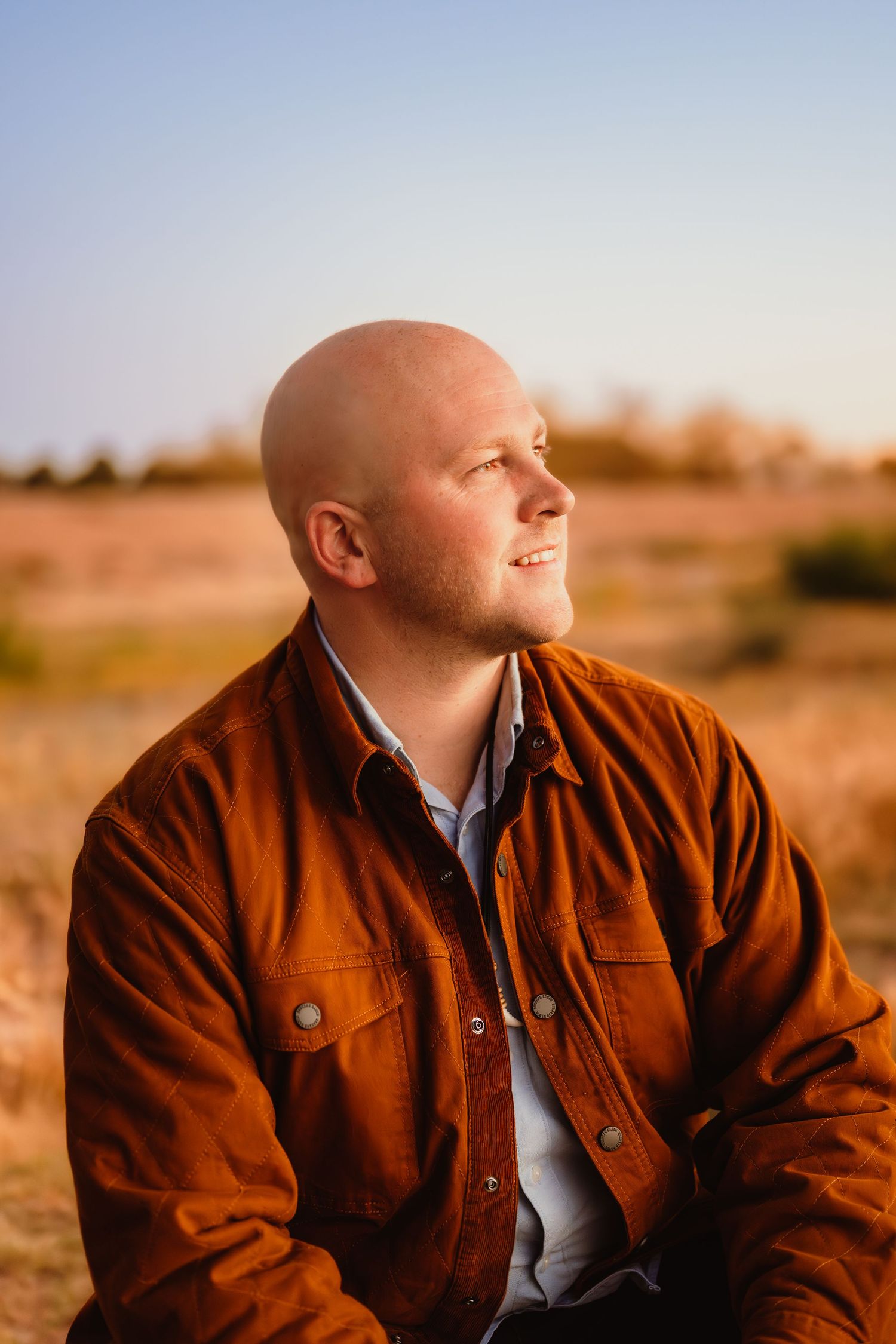 A person in a brown leather jacket gazes thoughtfully into the distance against a desert landscape at sunset.