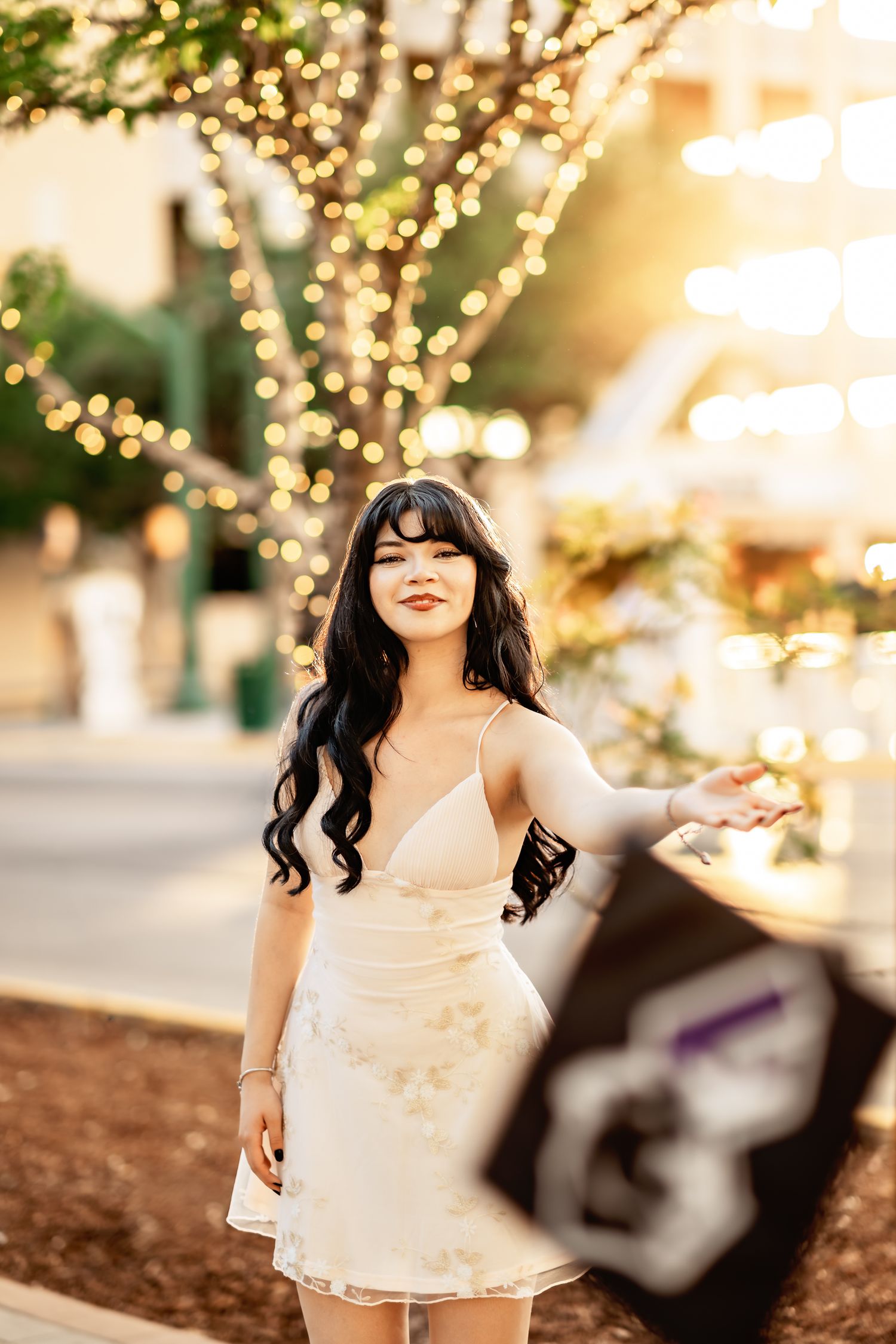 Young woman in white floral dress poses outdoors at dusk near a tree adorned with warm bokeh string lights.