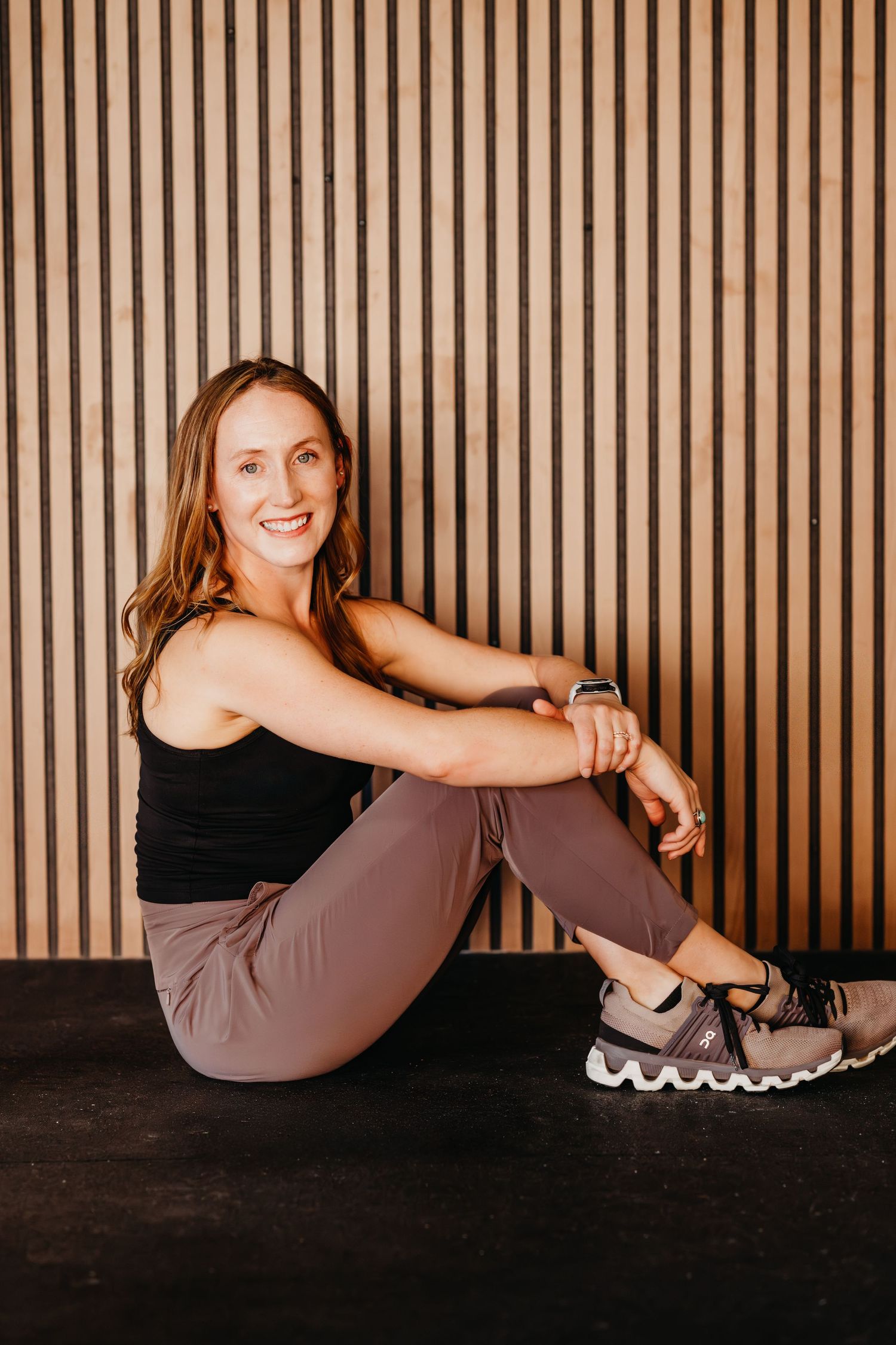 Person in athletic wear sits casually against wooden slat wall wearing black tank top, mauve pants and sneakers.