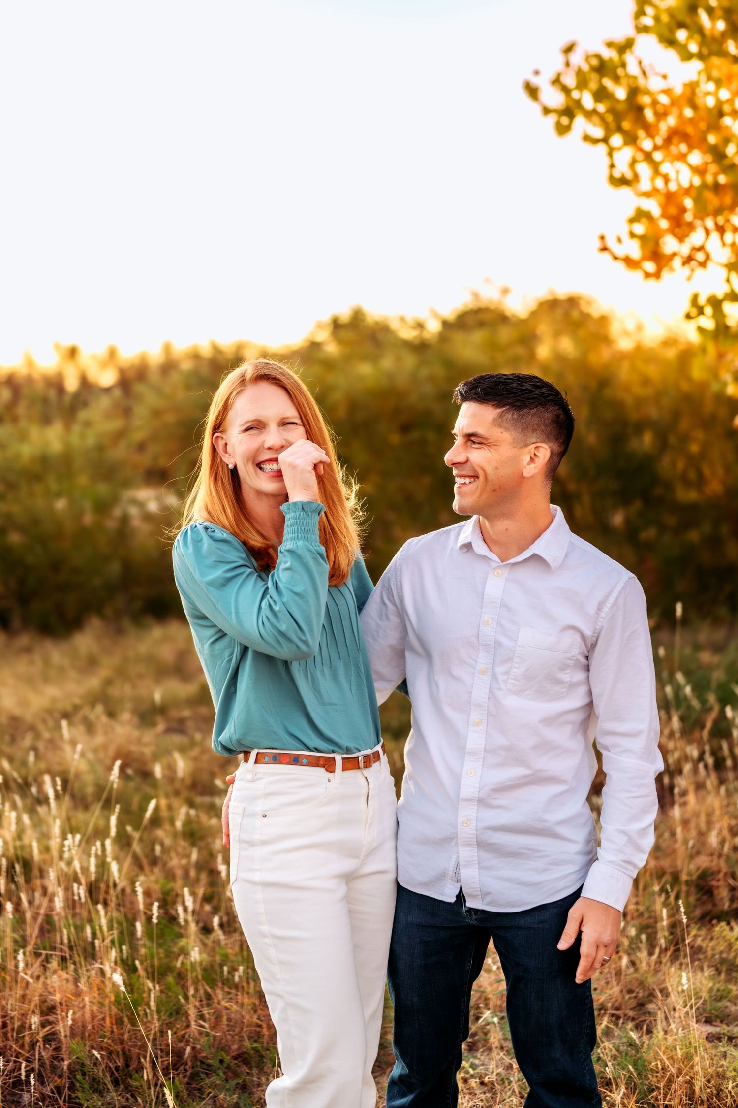 A couple laughs together in a field during golden hour wearing casual coordinated outfits.