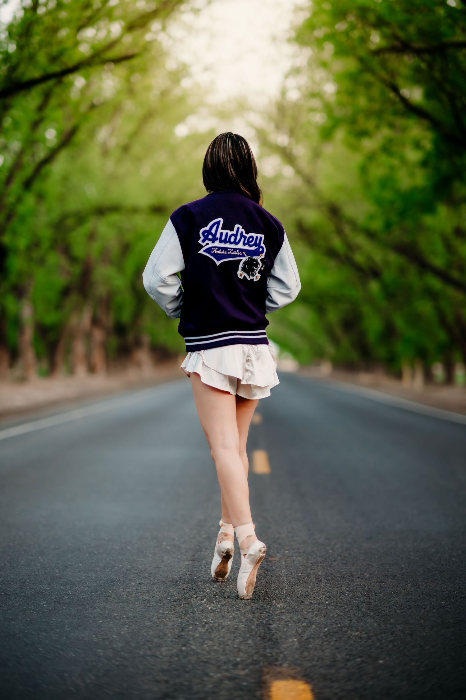 Ballet dancer in varsity jacket stands en pointe on empty tree-lined road wearing pointe shoes and shorts.