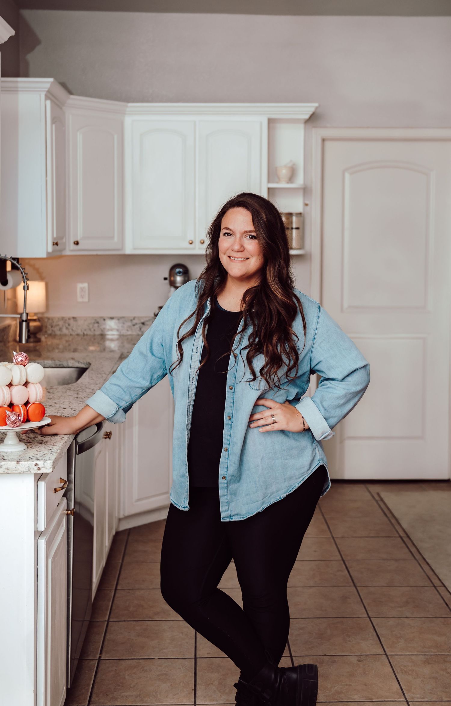 A person in a light blue denim shirt and black pants stands casually in a modern white kitchen with granite countertops.