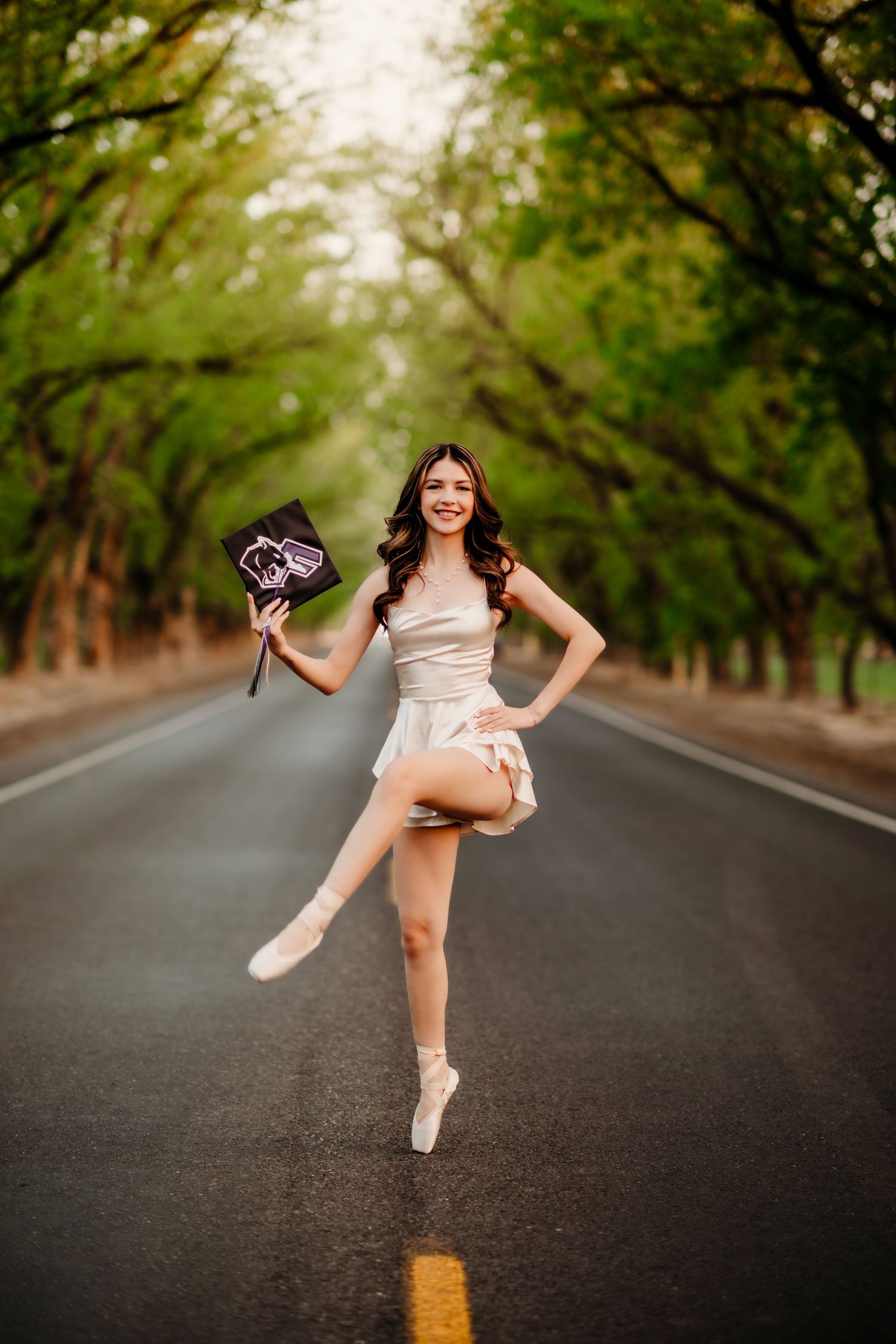 Ballet dancer in pointe shoes performs graceful poses on a tree-lined road during a golden hour photoshoot.