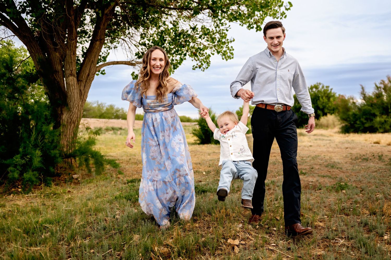 Family swinging toddler between them outdoors in green field, parents smiling, woman in blue floral dress, man in gray shirt.