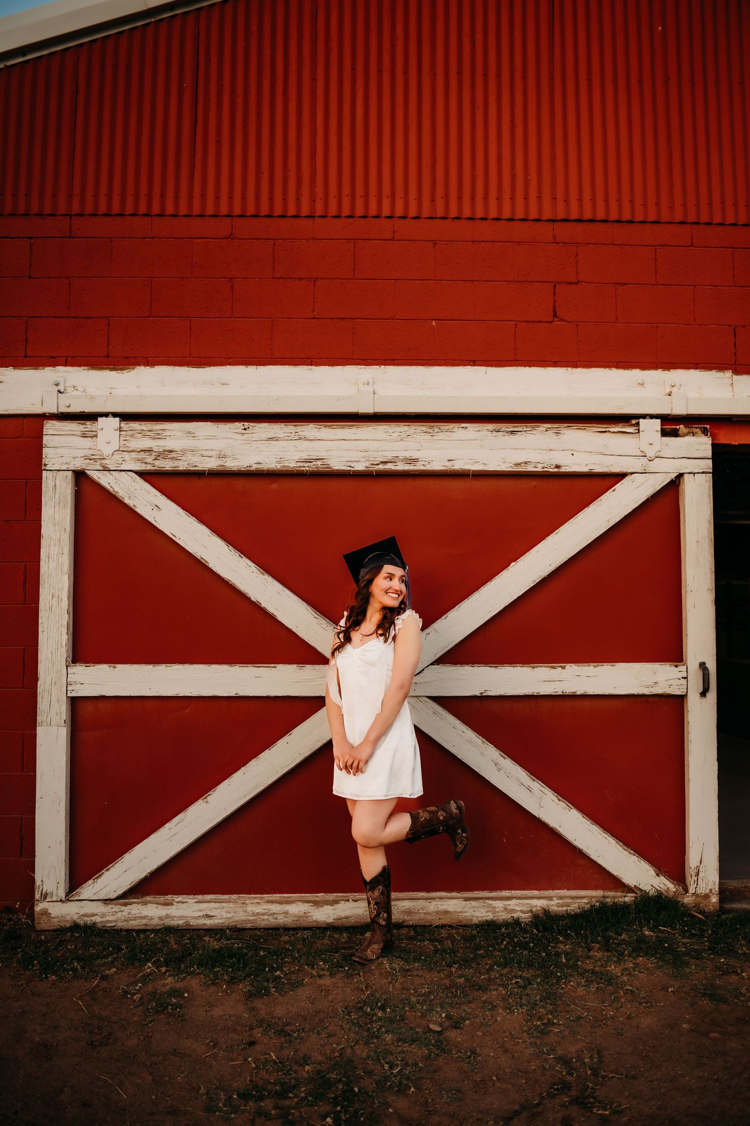Graduate in a white dress poses against a red barn door with white cross beams during sunset.