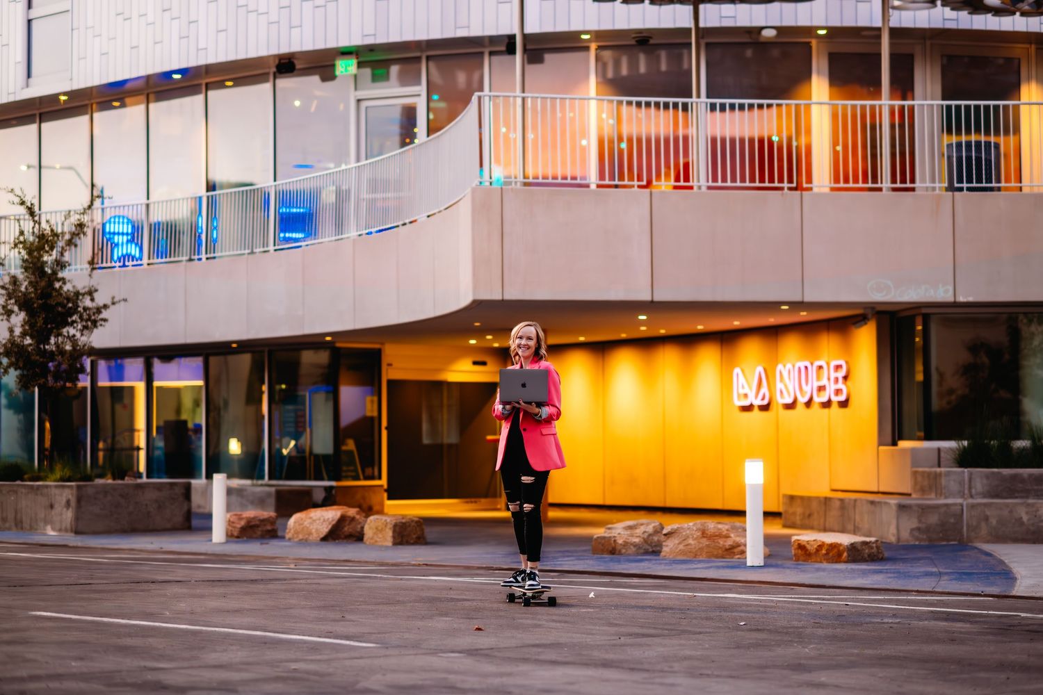 Skateboarder performs trick at dusk in front of illuminated building entrance with orange glow.