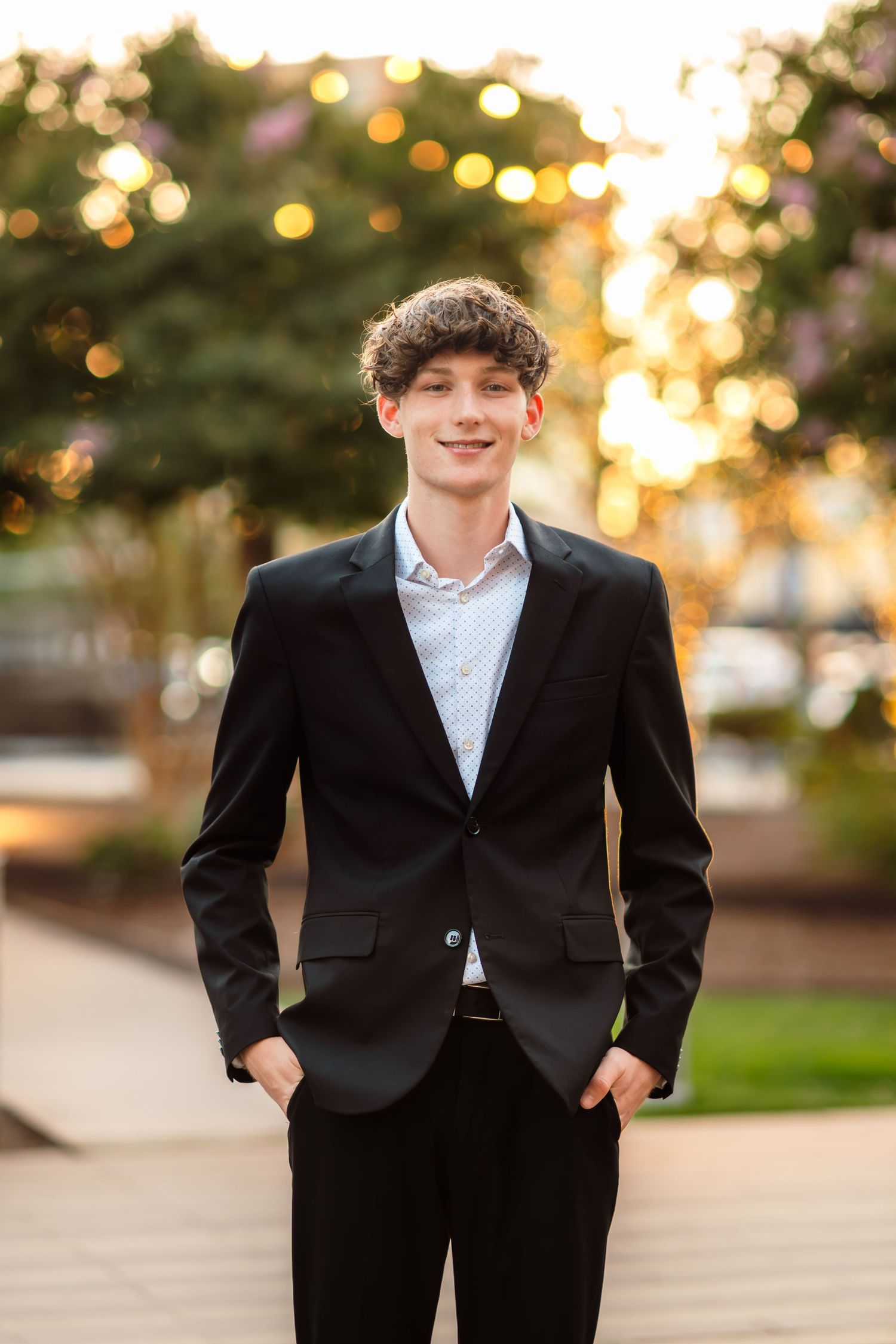 Figure in formal black suit standing on outdoor pathway during sunset.