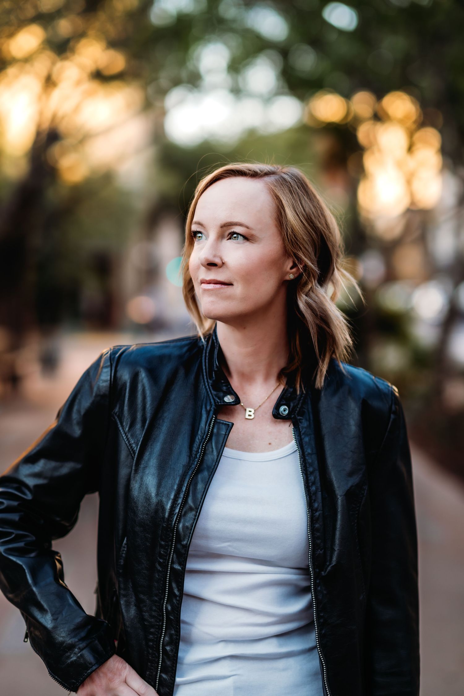 Woman in black leather jacket and white tank top poses outdoors at sunset with natural bokeh lighting effect.