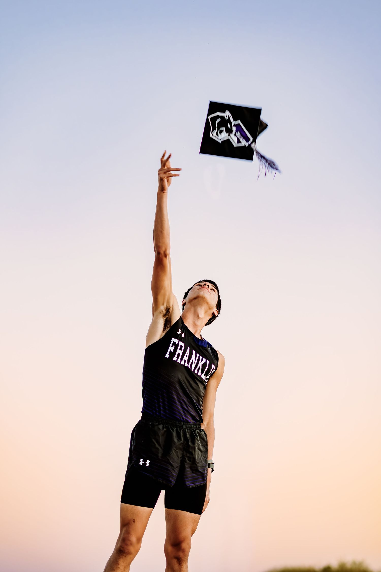 Athlete throws graduation cap in air against sunset sky.