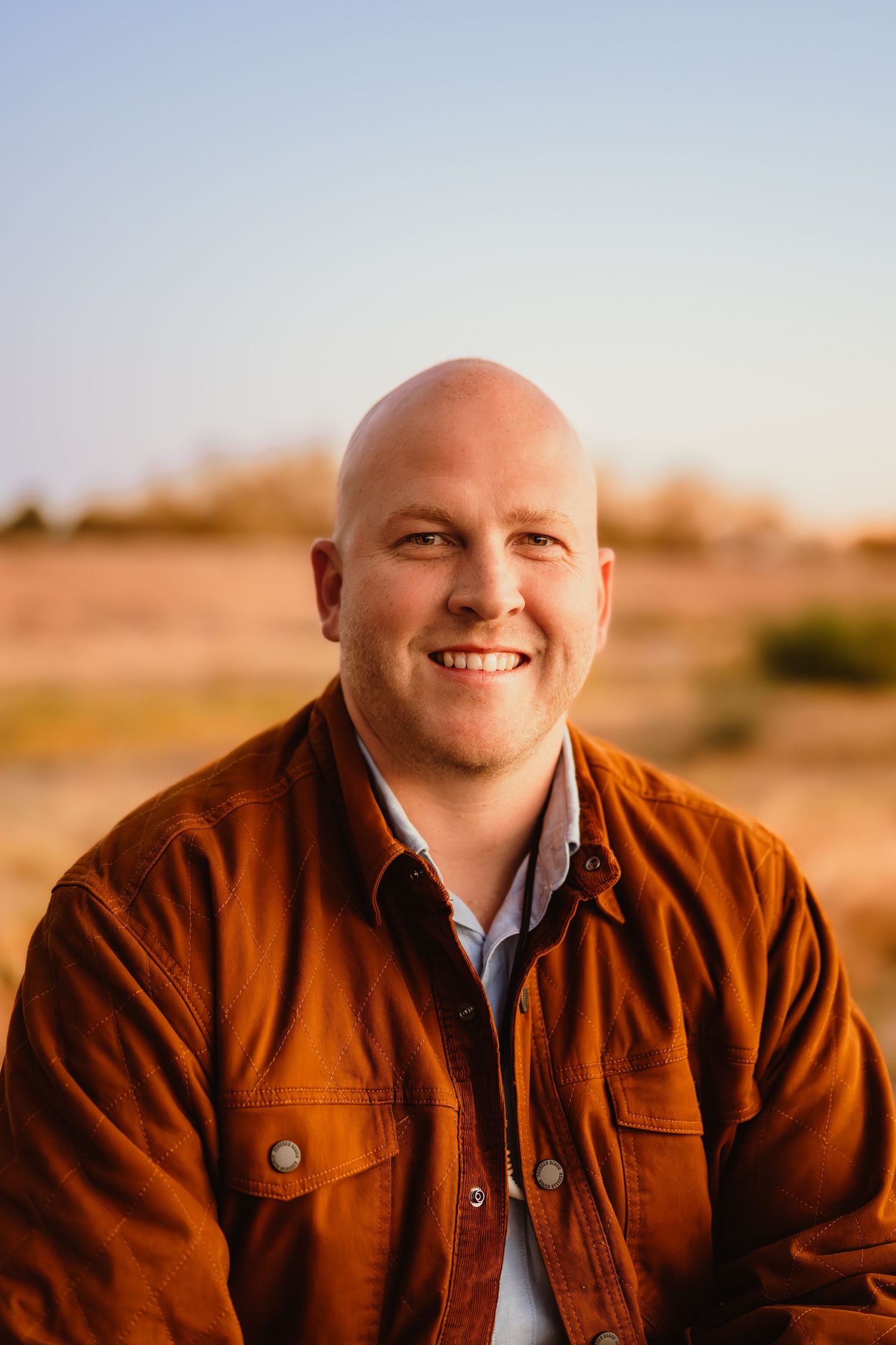 A person wearing a brown jacket smiles warmly at sunset in a desert landscape setting.