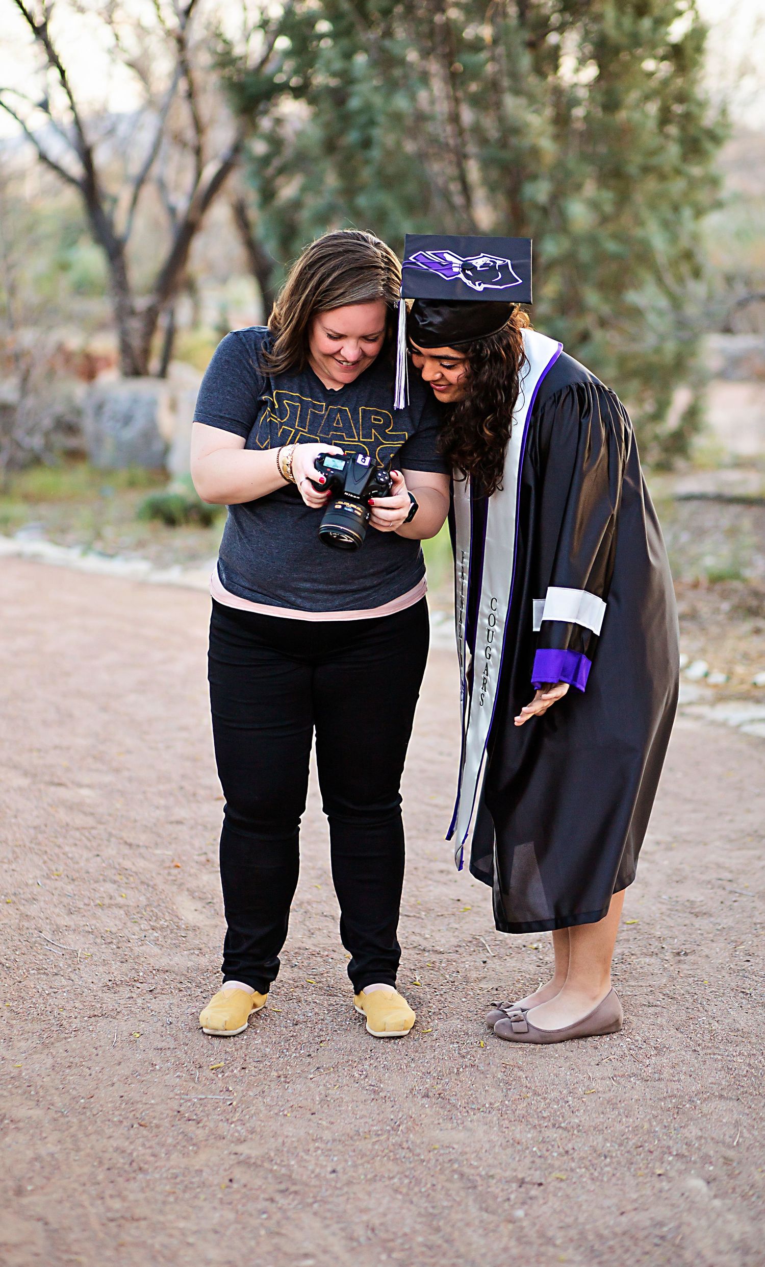 Two people look at a camera display while one wears graduation regalia outdoors.