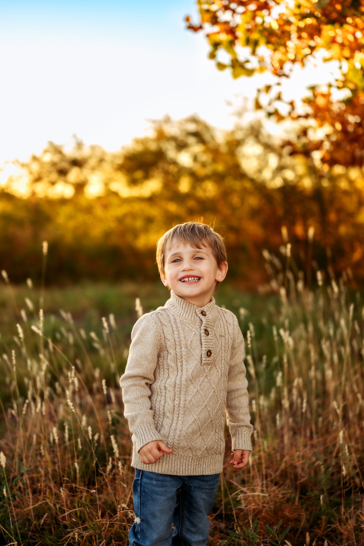 A child wearing a beige cable knit sweater smiles joyfully in a golden autumn field at sunset.