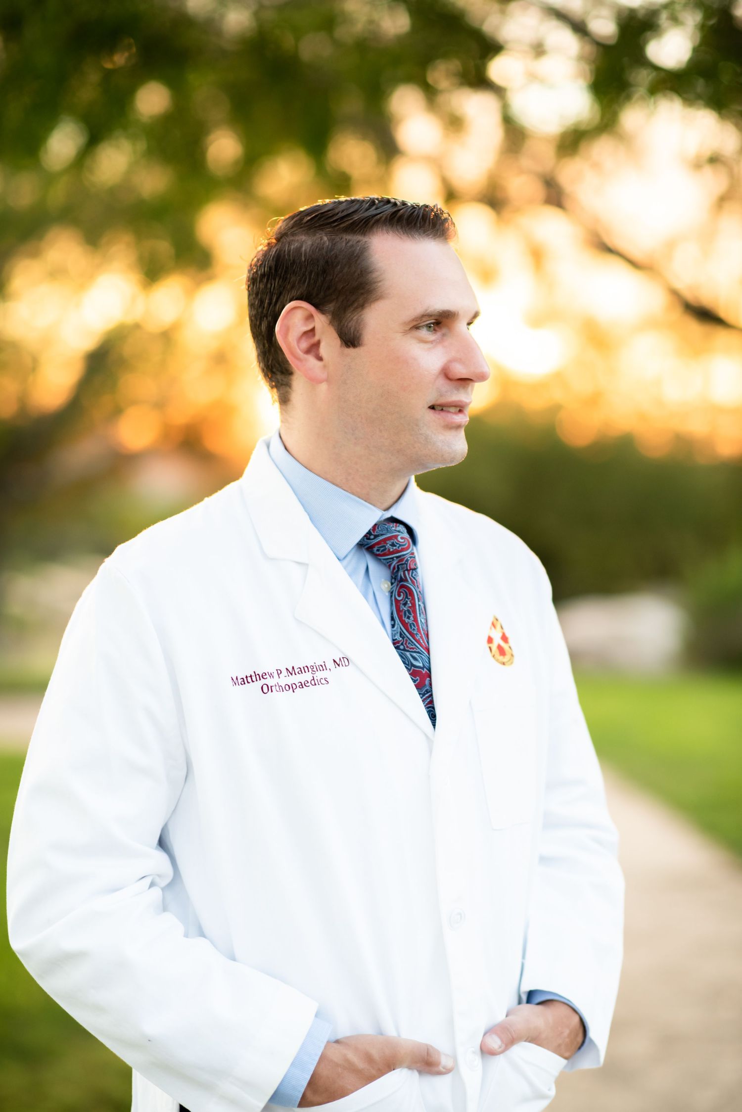Medical professional in white coat and tie stands outdoors during golden hour lighting.