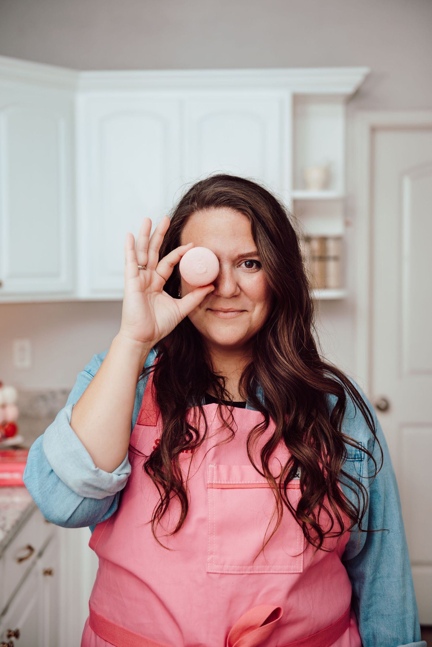 Person in pink shirt and denim jacket making playful gestures with hands in front of their face in a bright kitchen.