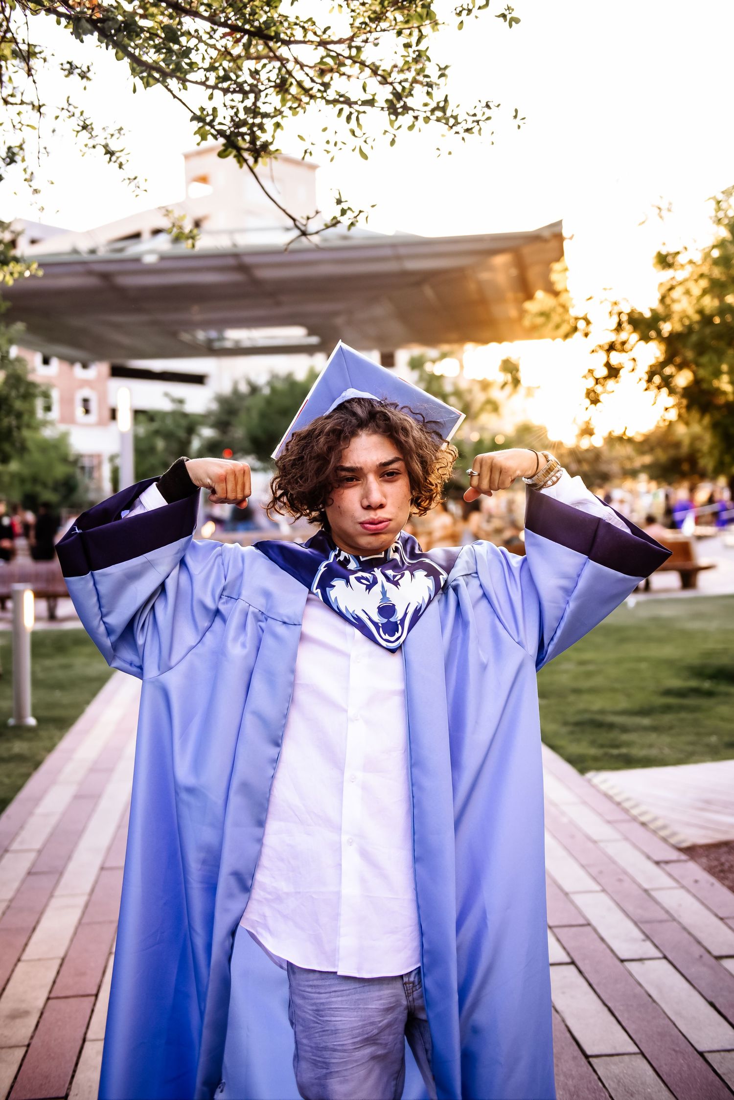A graduate in a light blue graduation gown poses joyfully with outstretched arms at sunset on a brick walkway.