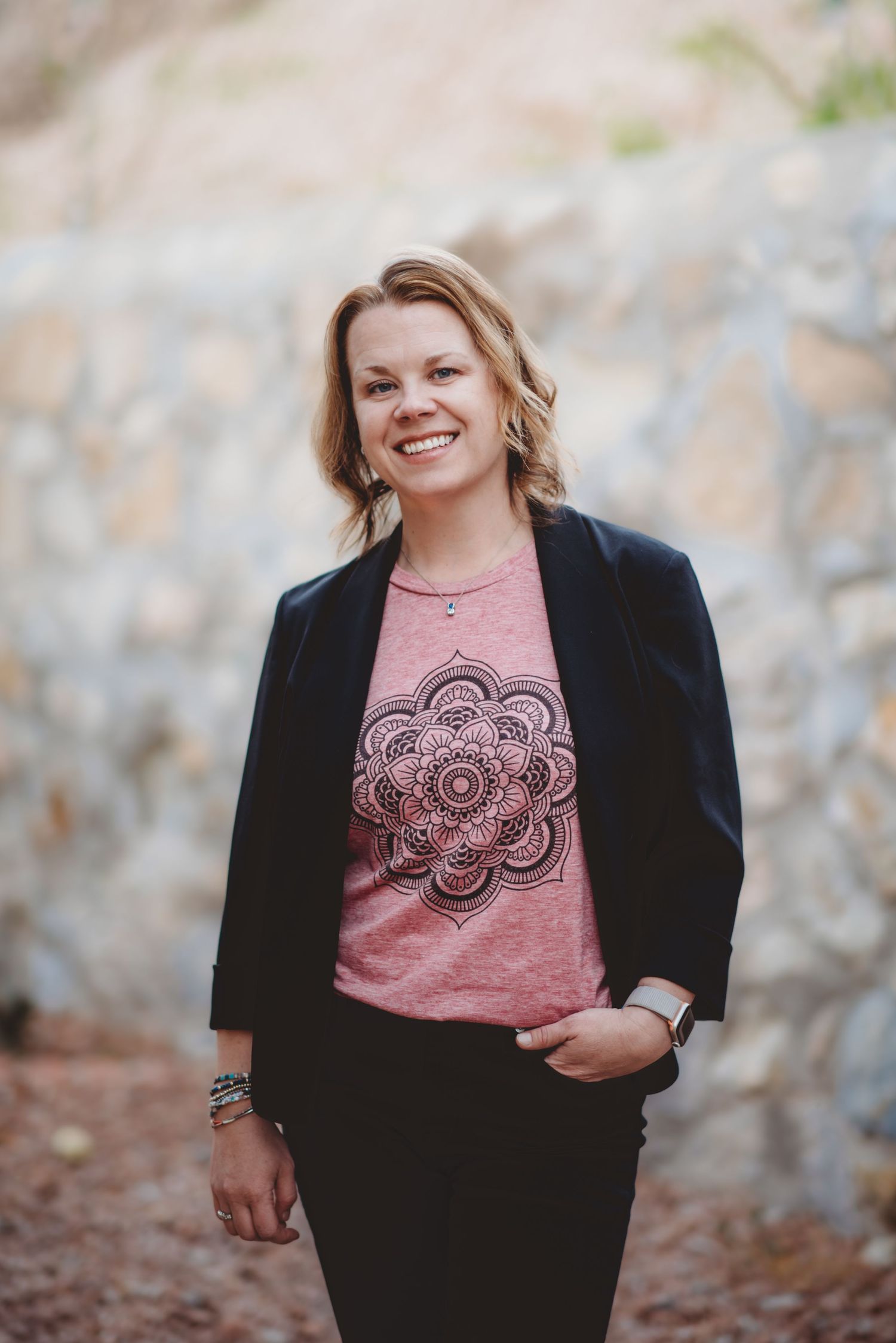 A person wearing a pink t-shirt with mandala design and black cardigan poses outdoors with a blurred background.