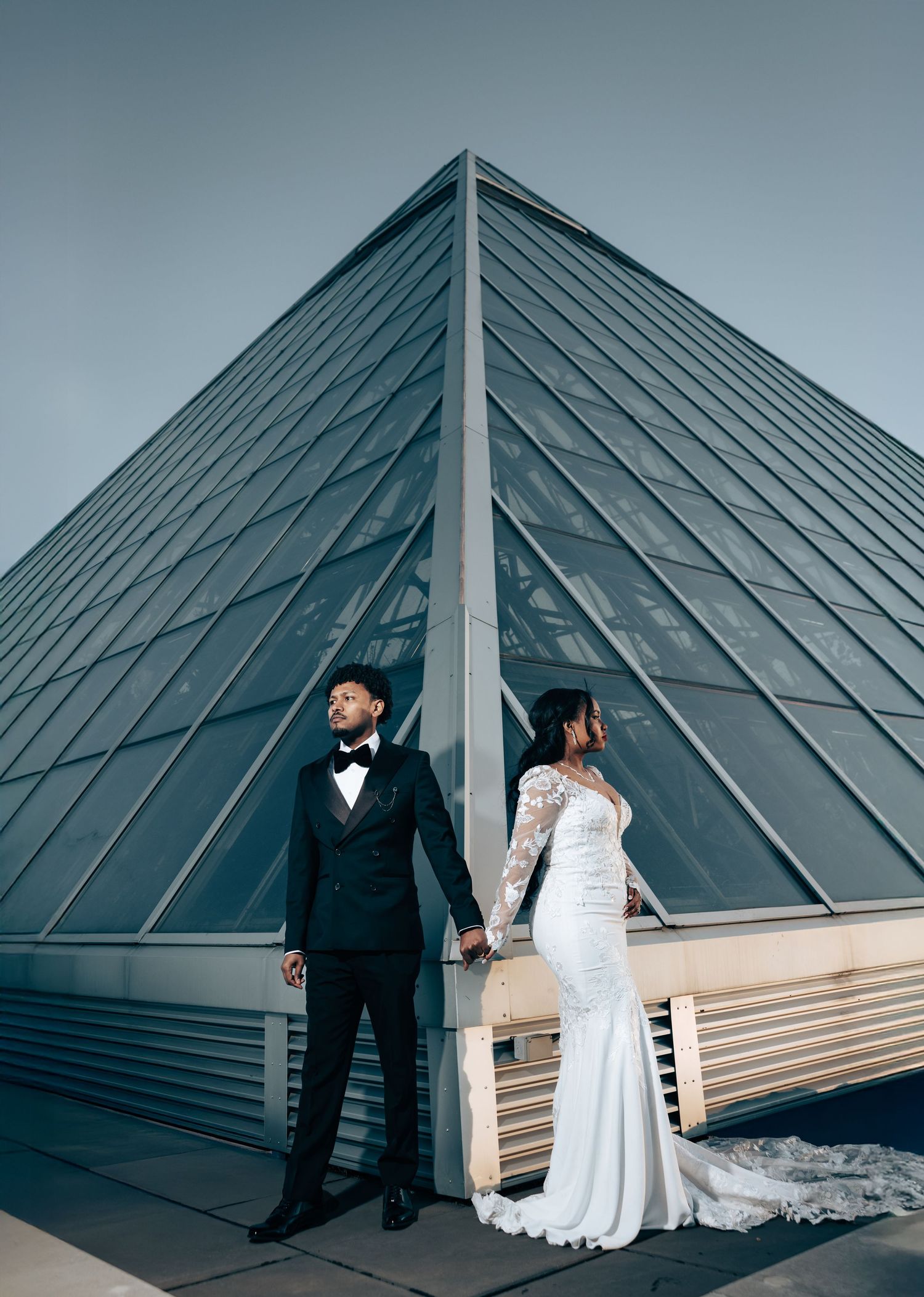 Wedding couple poses against modern glass pyramid architecture in dramatic composition.