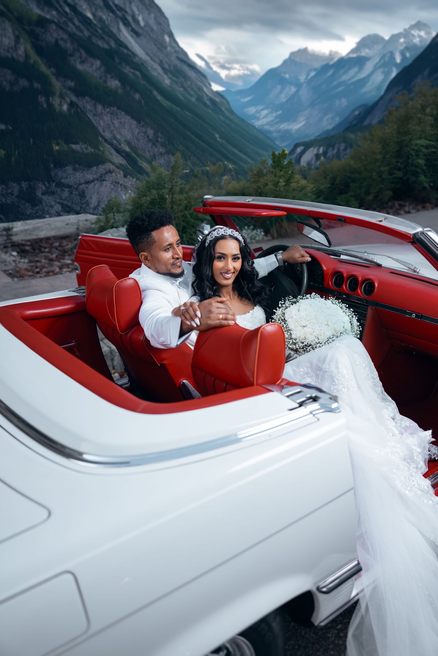 Couple in white vintage convertible car against dramatic mountain landscape backdrop.